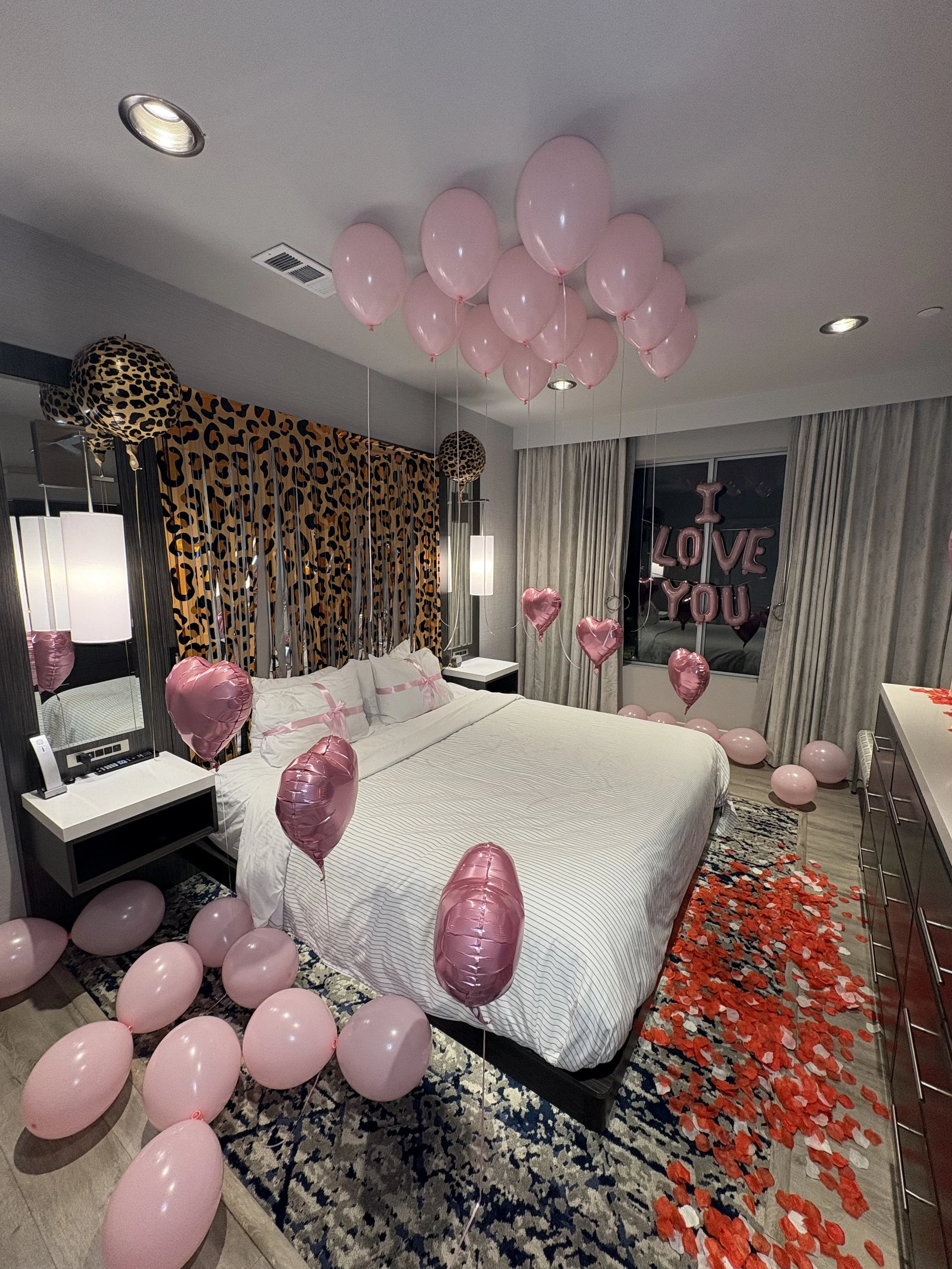 Bedroom decorated with pink balloons, heart-shaped balloons, LOVE YOU letter balloons, leopard-print curtains, and rose petals.