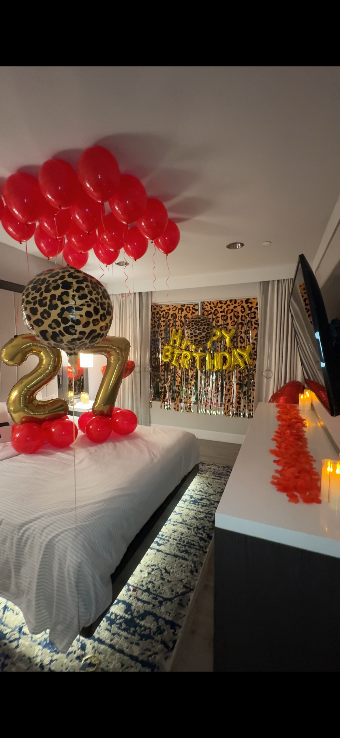 Hotel bedroom decorated for a 27th birthday celebration with red balloons, leopard-print balloons and backdrop, gold “27” balloons, and “Happy Birthday” lettering.