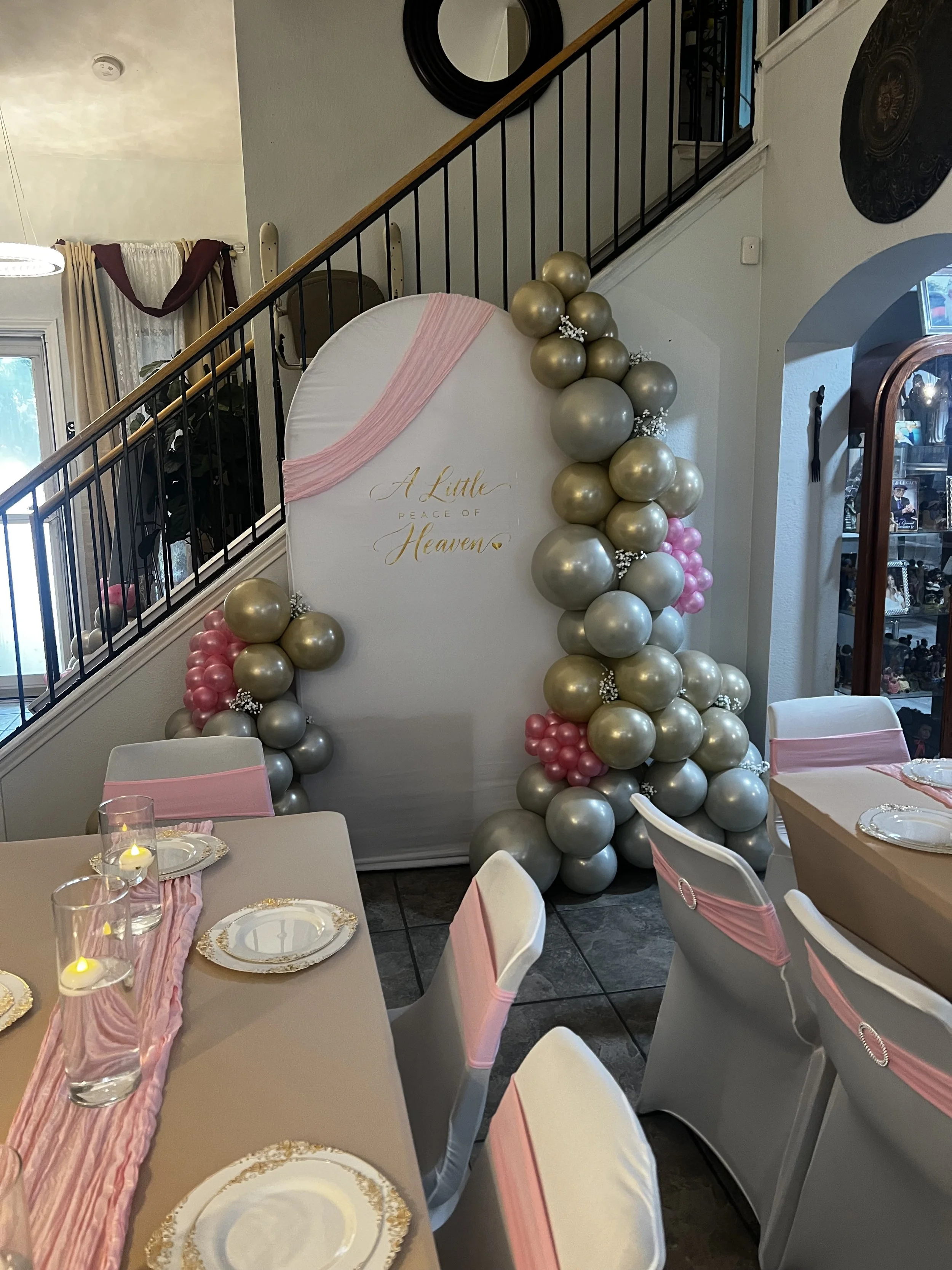 Elegant indoor celebration setup with pink and gold balloon garlands, a memorial sign reading “A Little Peace of Heaven,” and tables set with gold-trimmed plates and candles.
