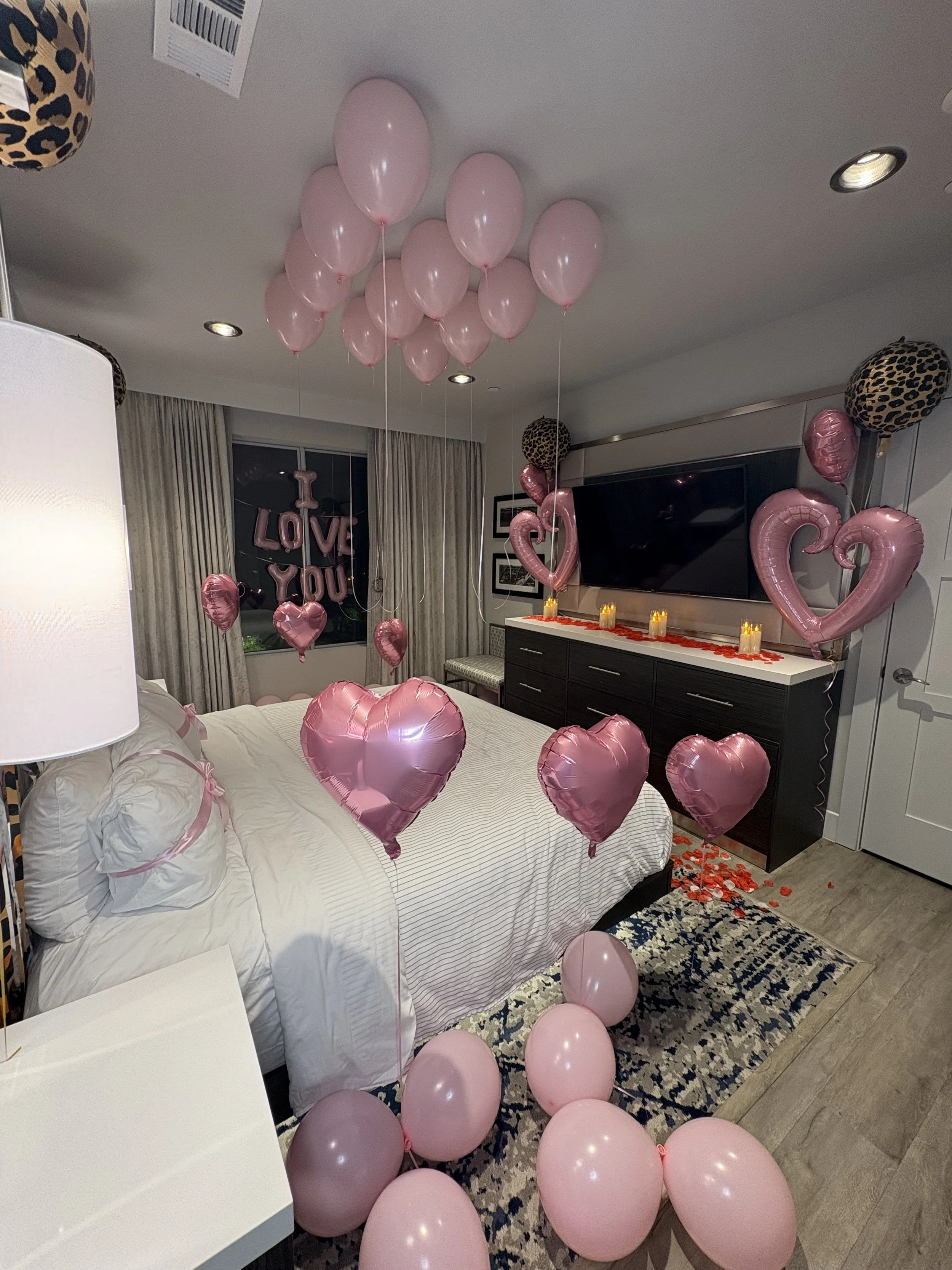 Bedroom decorated for a romantic celebration with pink heart balloons, floating balloons, rose petals, candles, and “LOVE YOU” balloon letters.