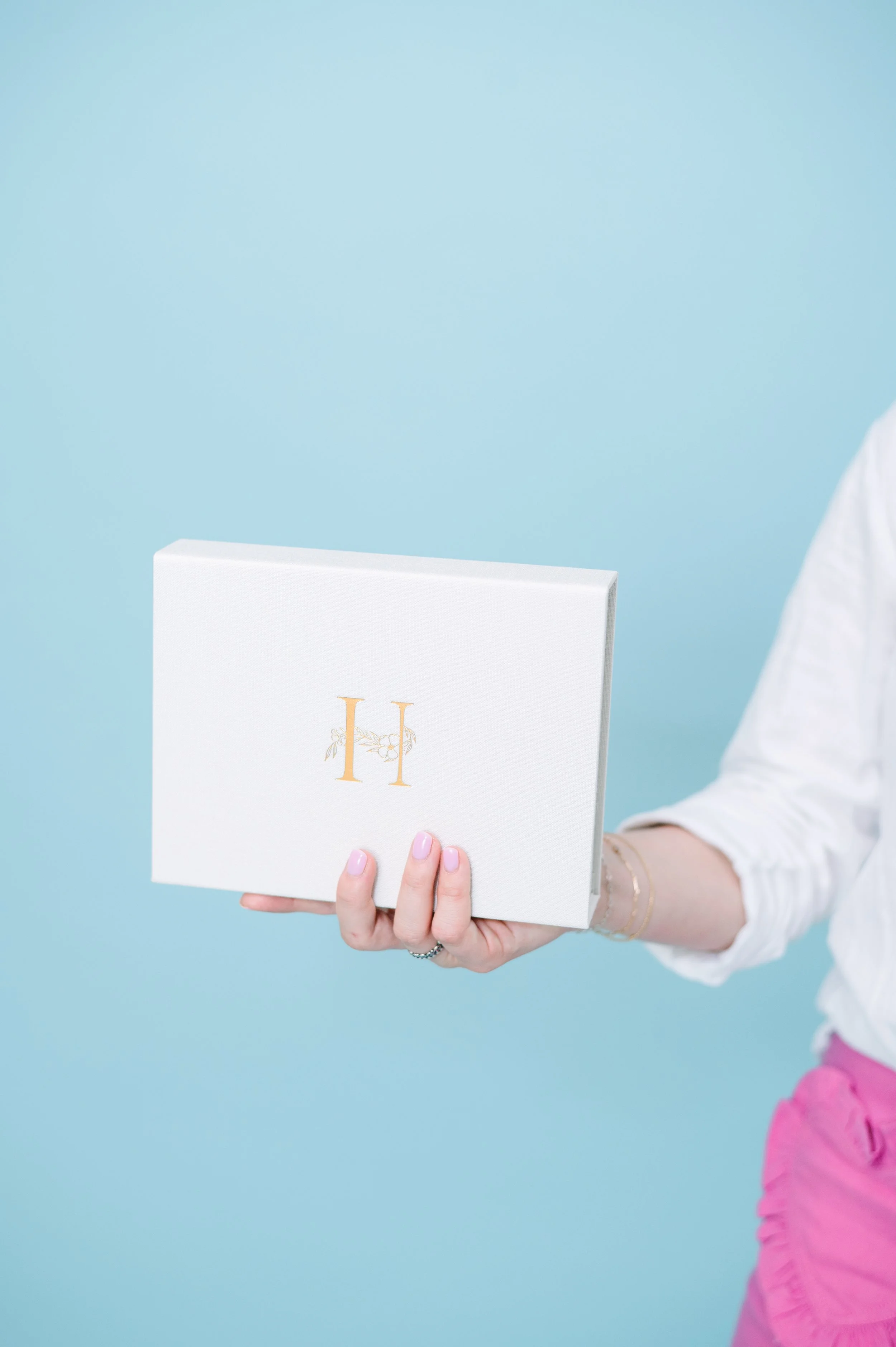 A person holding a white box with gold lettering and a floral illustration against a light blue background.
