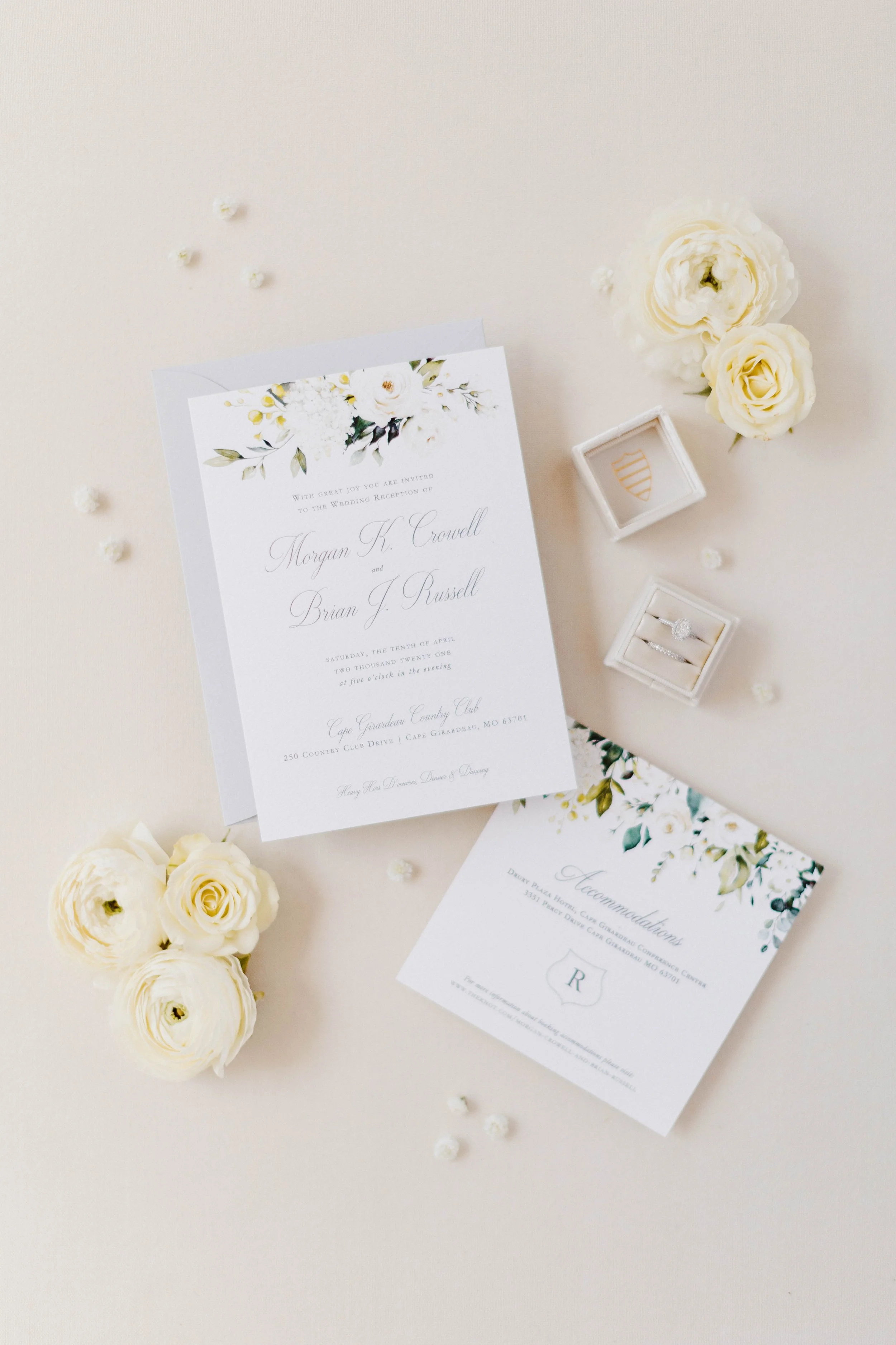 Wedding invitation card with floral decorations, surrounded by white flowers and engagement rings in a box, on a cream-colored background.