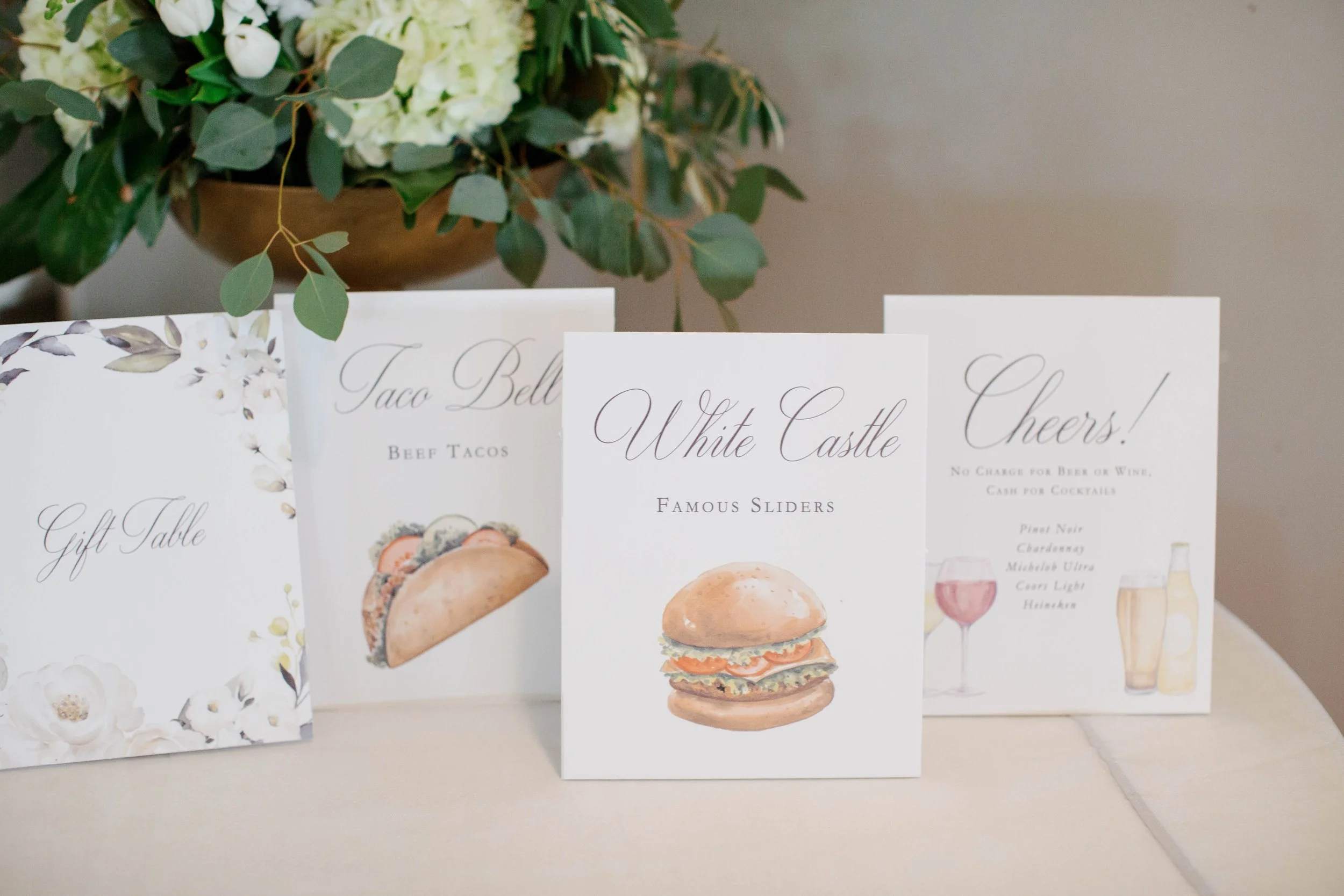 Display of menus for catering or a food event with various food and drink options, adorned with a flower arrangement in the background.