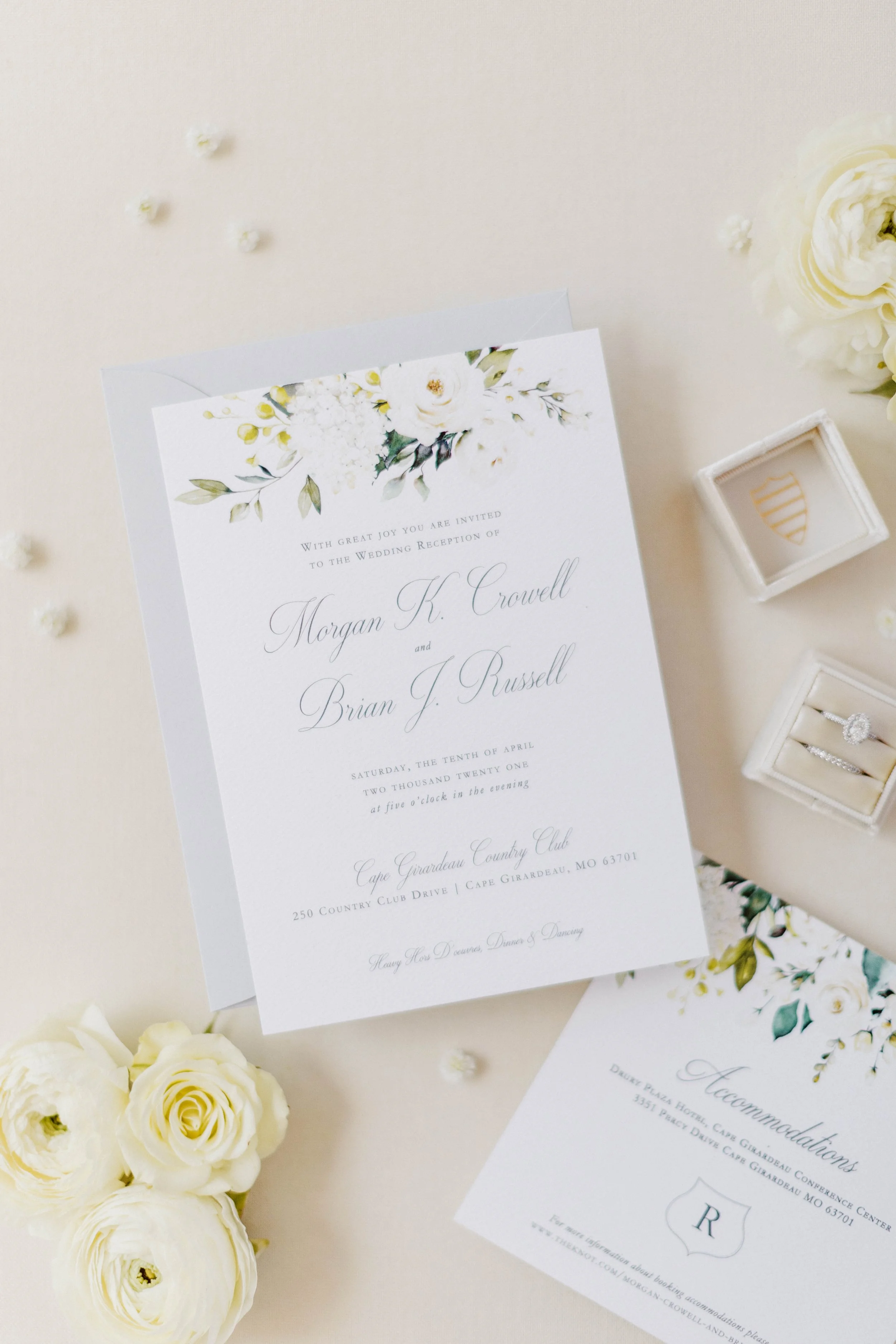 Elegant wedding invitation with floral design, surrounded by white roses and flowers, along with a small white ring box and a card of congratulations.
