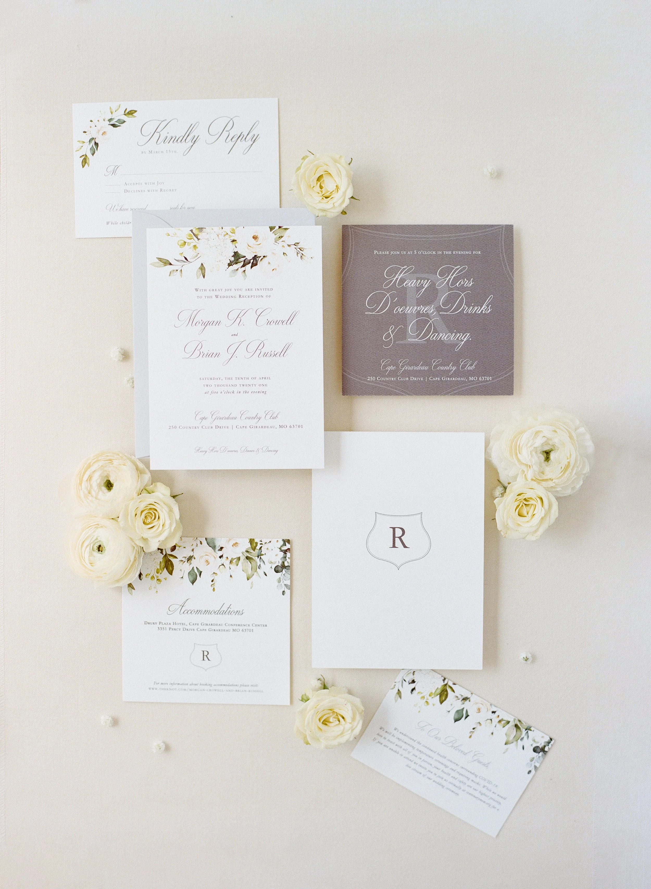 A flat lay of wedding invitations, RSVP card, accommodation details, and event information, accompanied by white roses and small white decorative balls, on a light background.