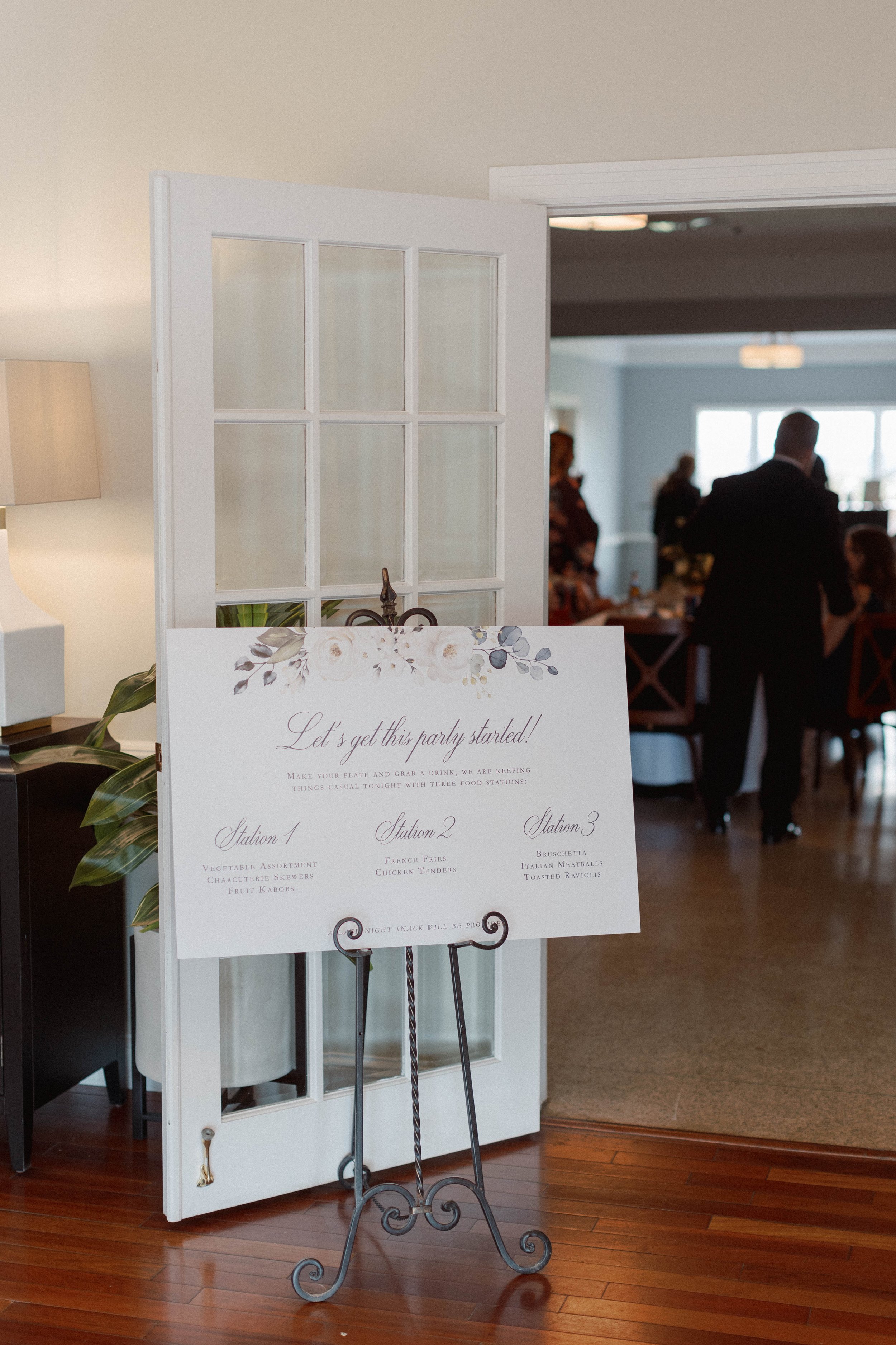 Event sign on easel with food station details at a gathering, partially visible with people in the background in a decorated indoor venue.