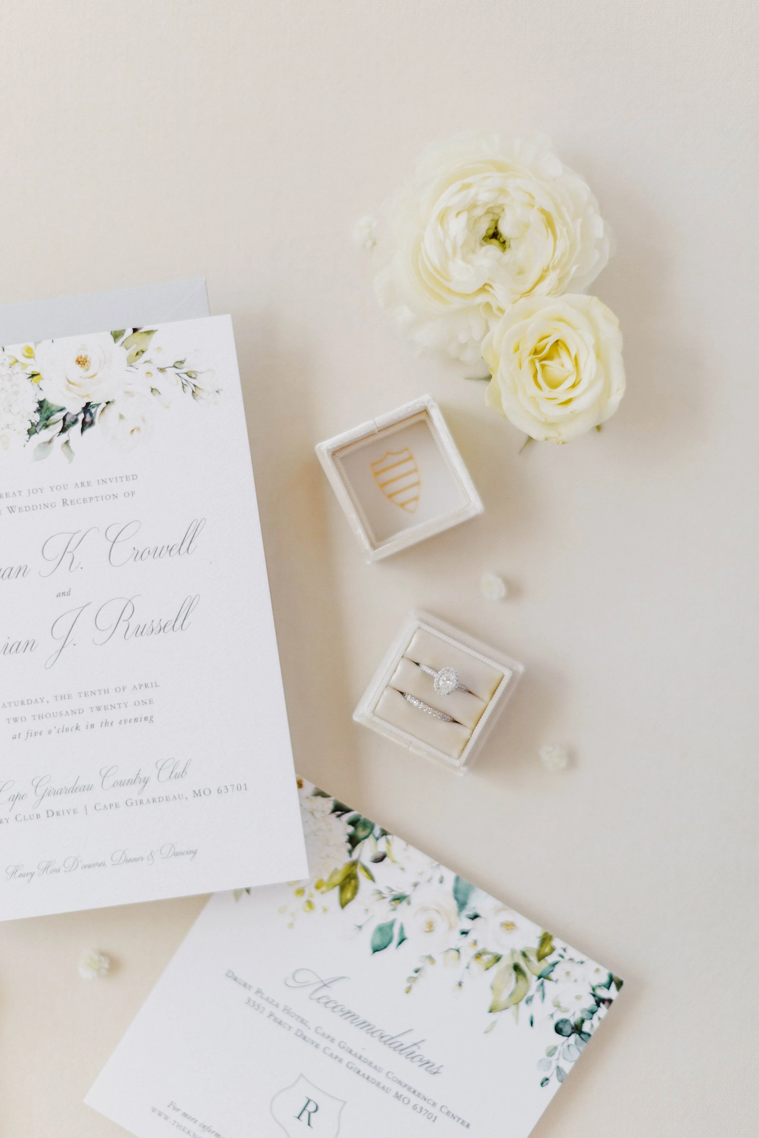 A wedding invitation card with floral decoration, a small box with a ring, white roses, and decorative items on a light-colored surface.