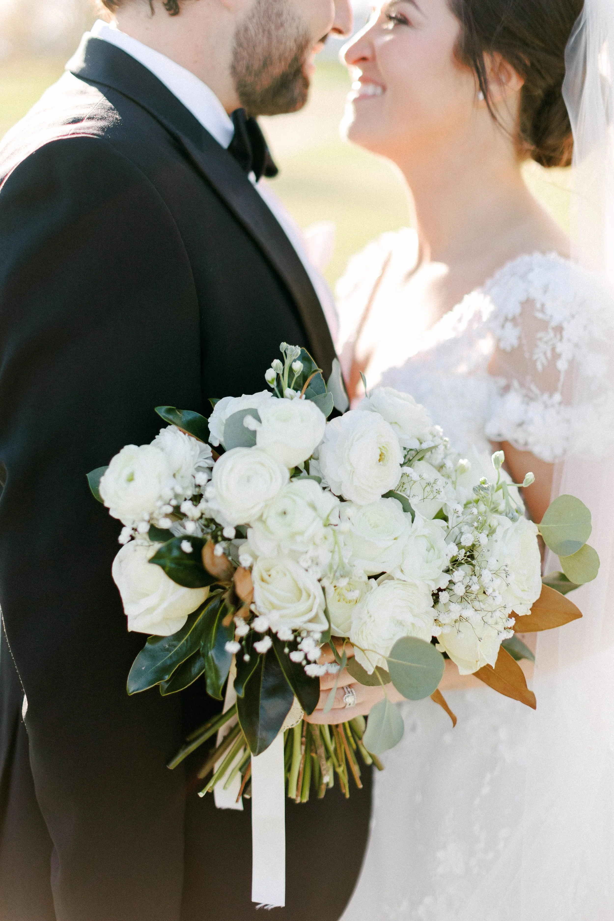 Close-up of a bride and groom on their wedding day, smiling at each other, with the bride holding a bouquet of white flowers.