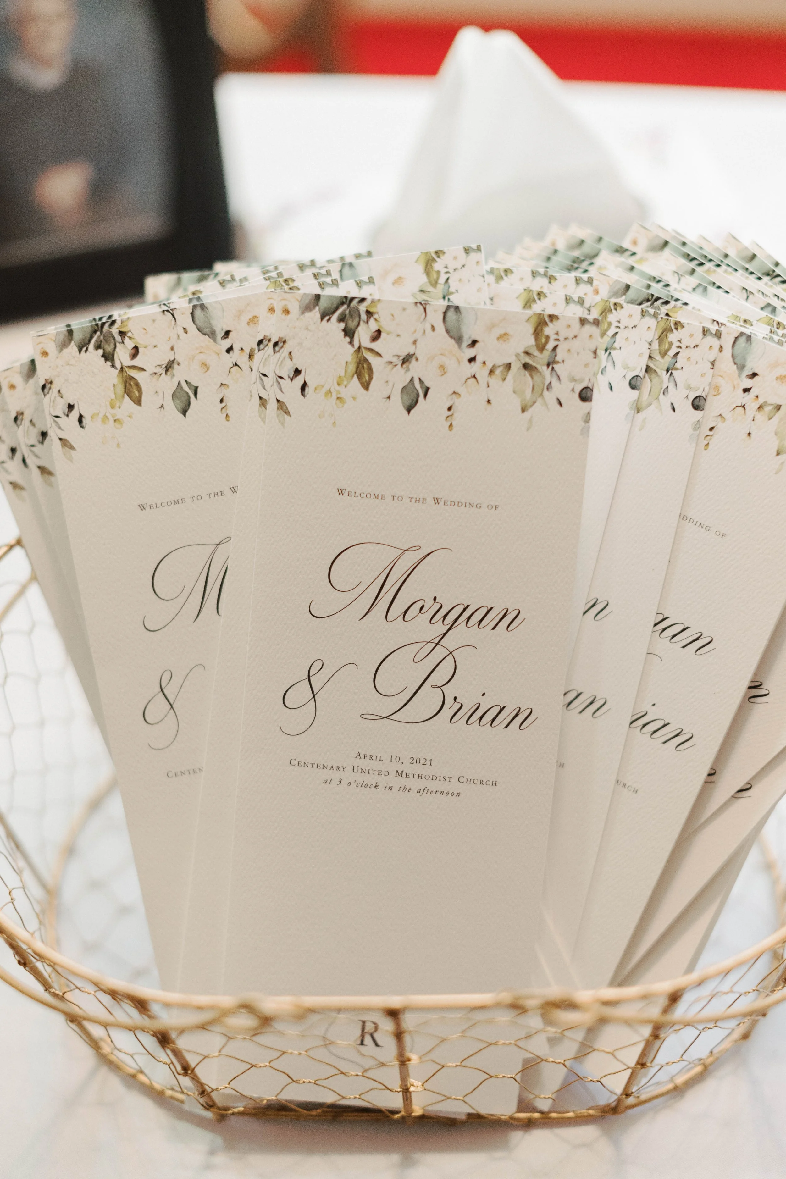 A set of wedding programs with floral designs and elegant fonts, placed inside a wire basket.