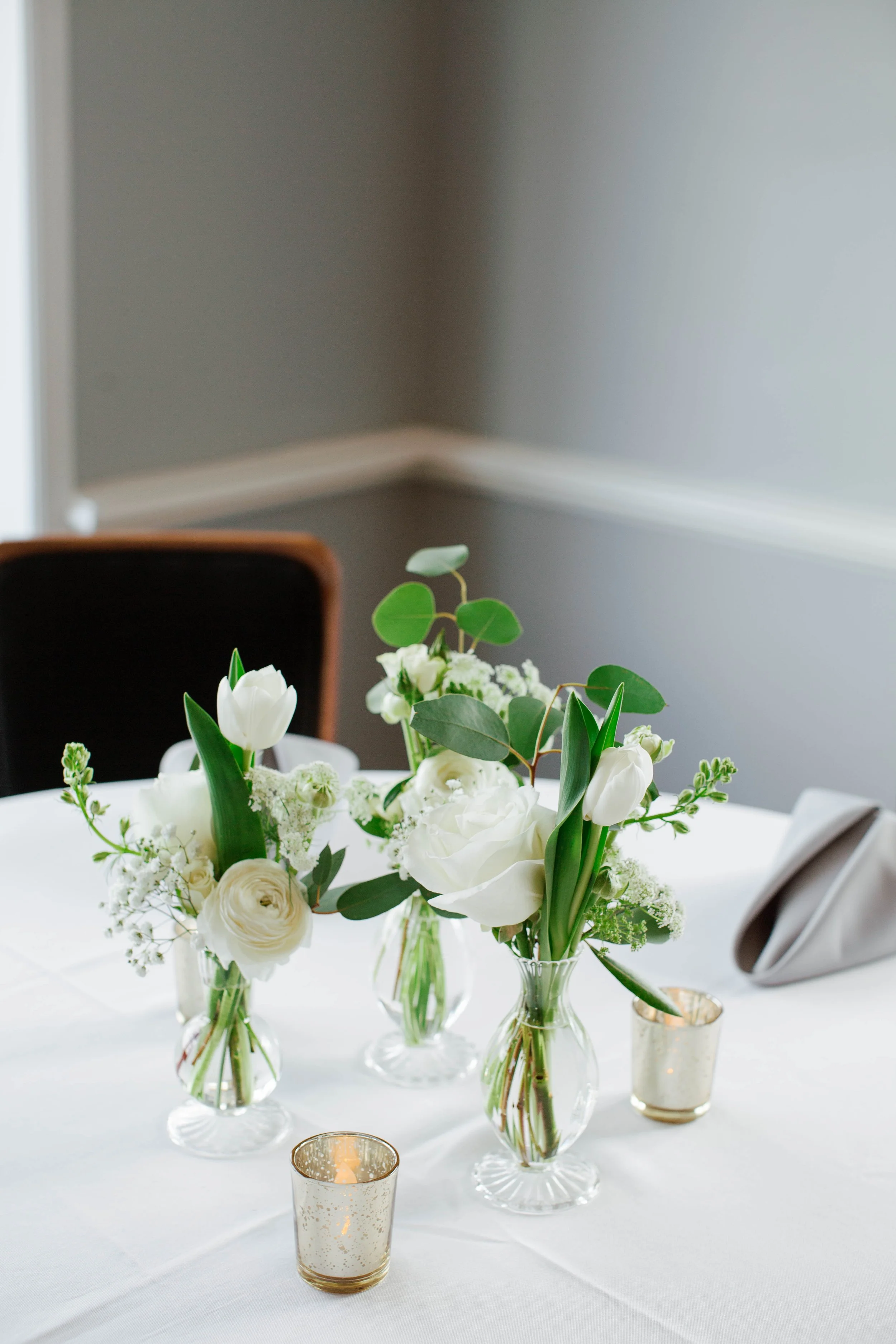 A table decorated with several small glass vases filled with white and green flowers, including tulips and greenery, and gold and glass candle holders.