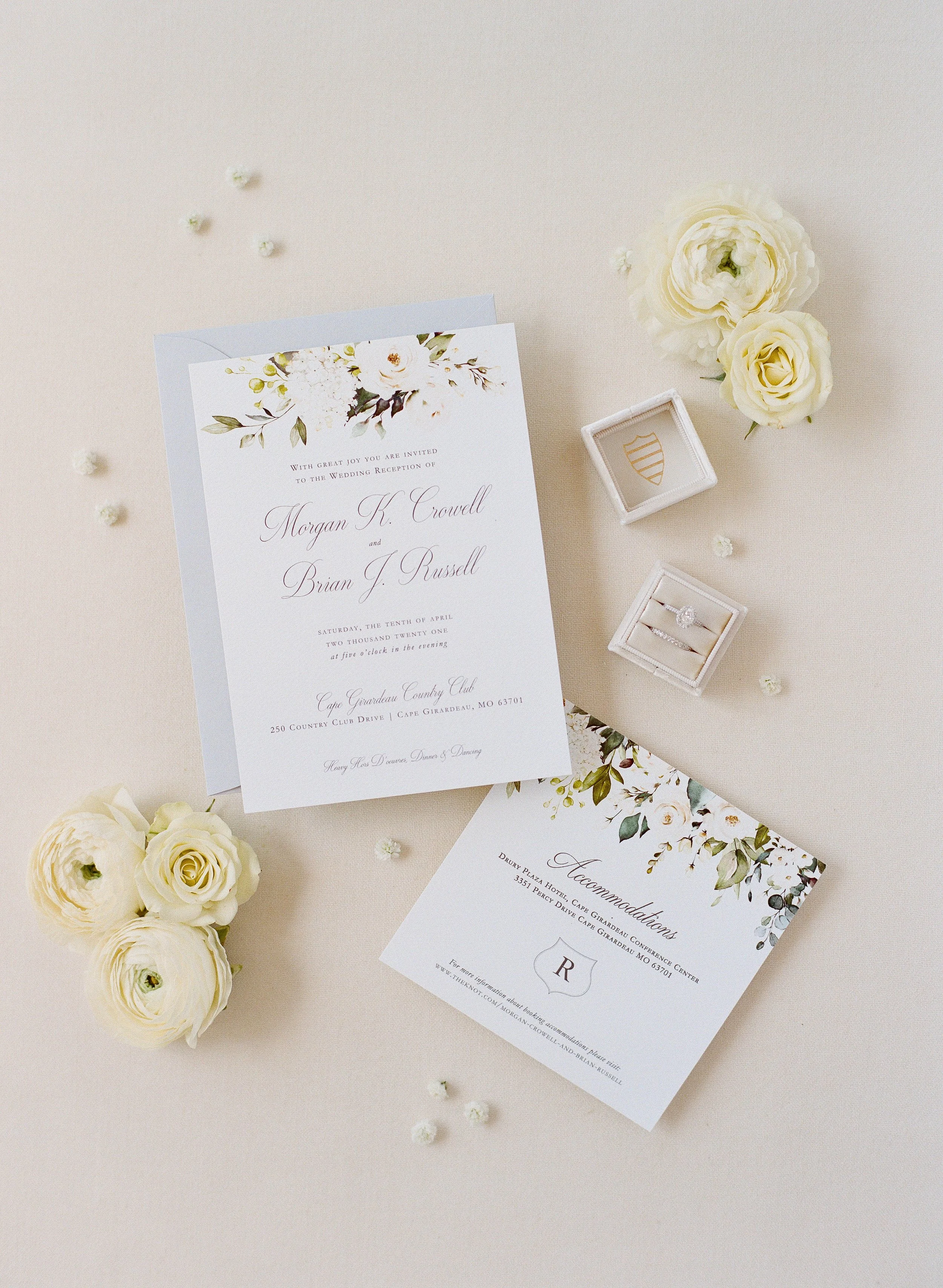 A wedding invitation with floral design on white card, surrounded by white roses and small flower petals, on a light background.