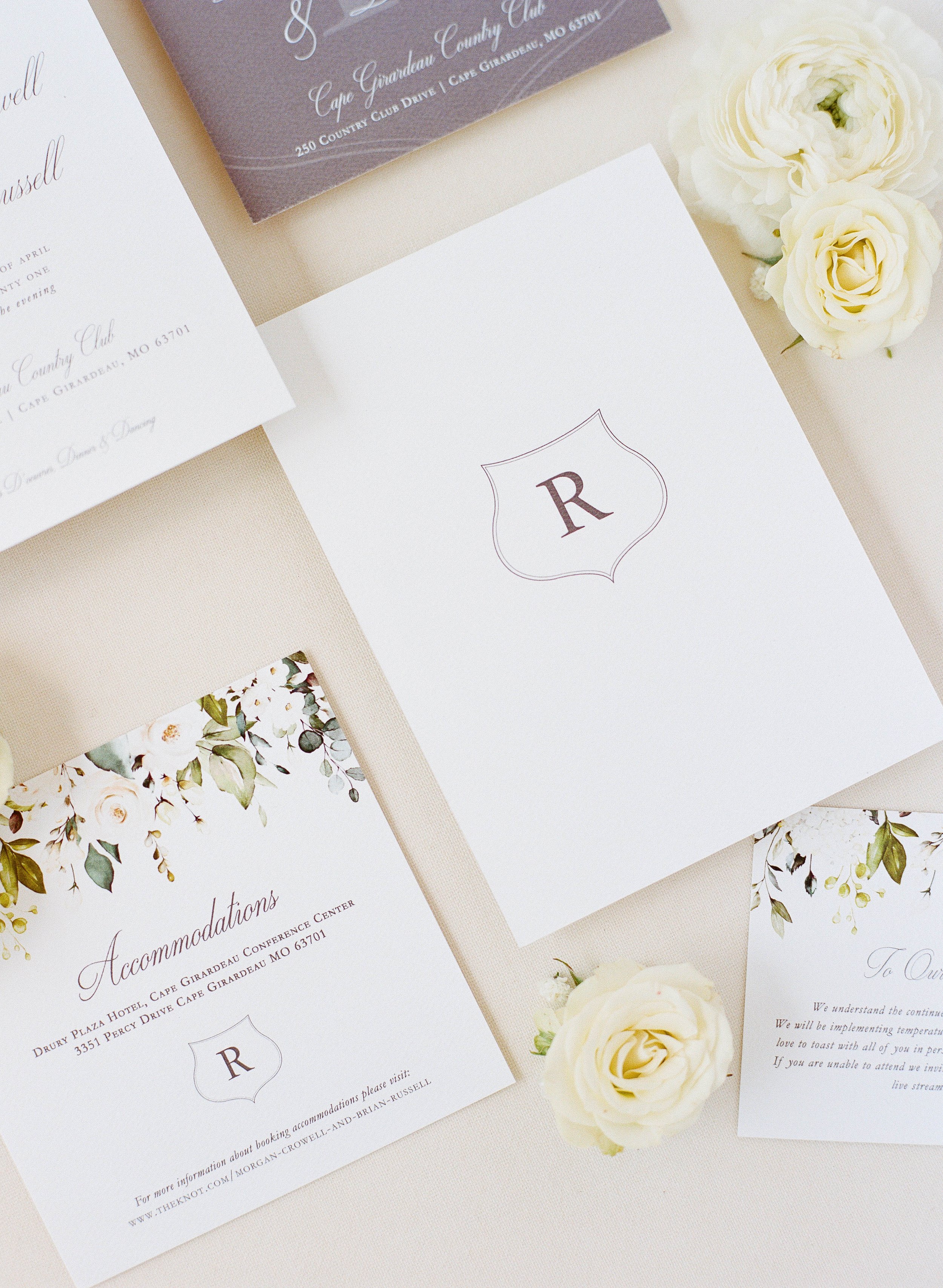 Wedding invitation cards with a monogram 'R', white roses, and floral designs on a beige background.