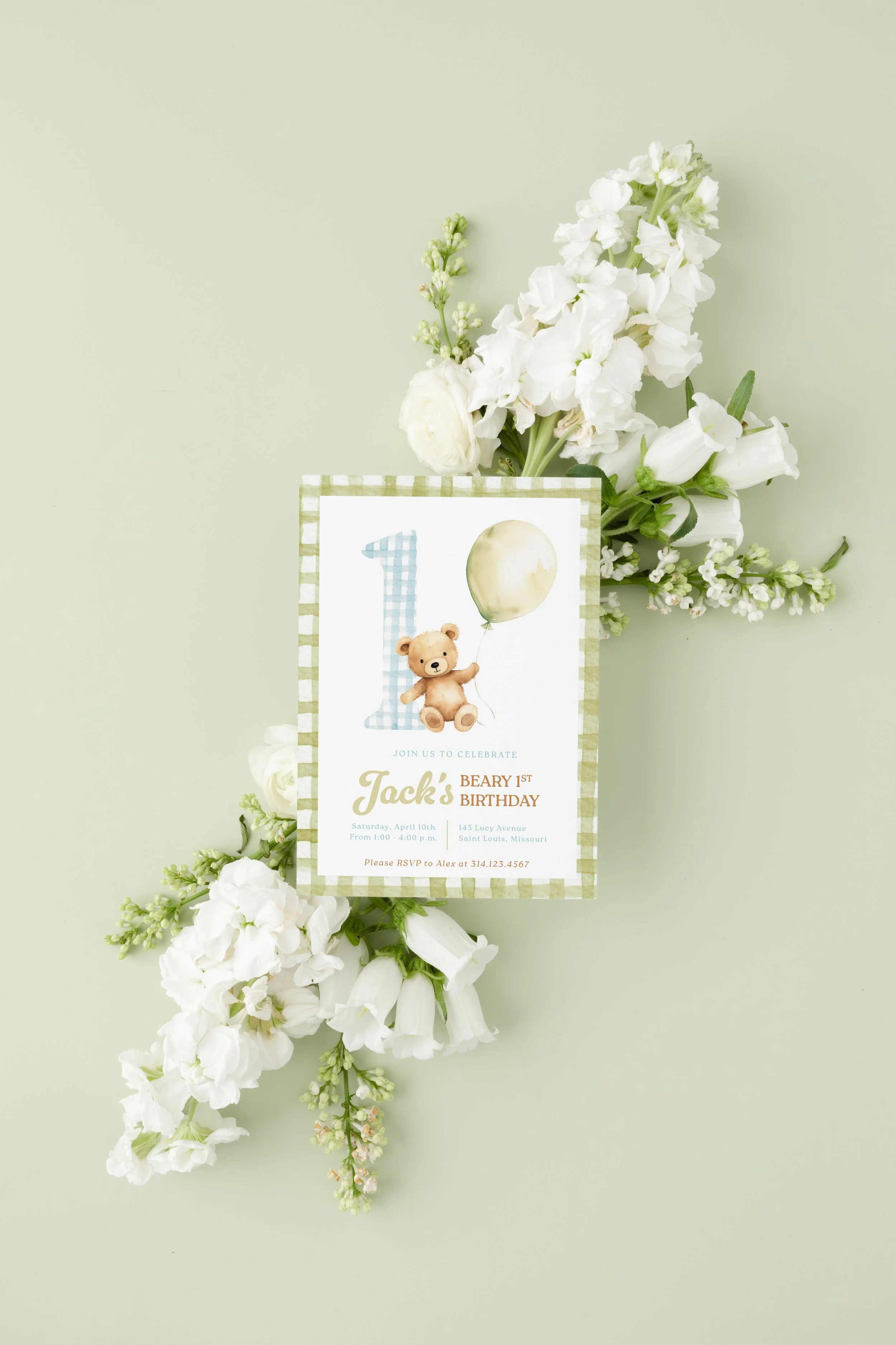 First birthday party invitation with a teddy bear holding a balloon, surrounded by white flowers on a light green background.