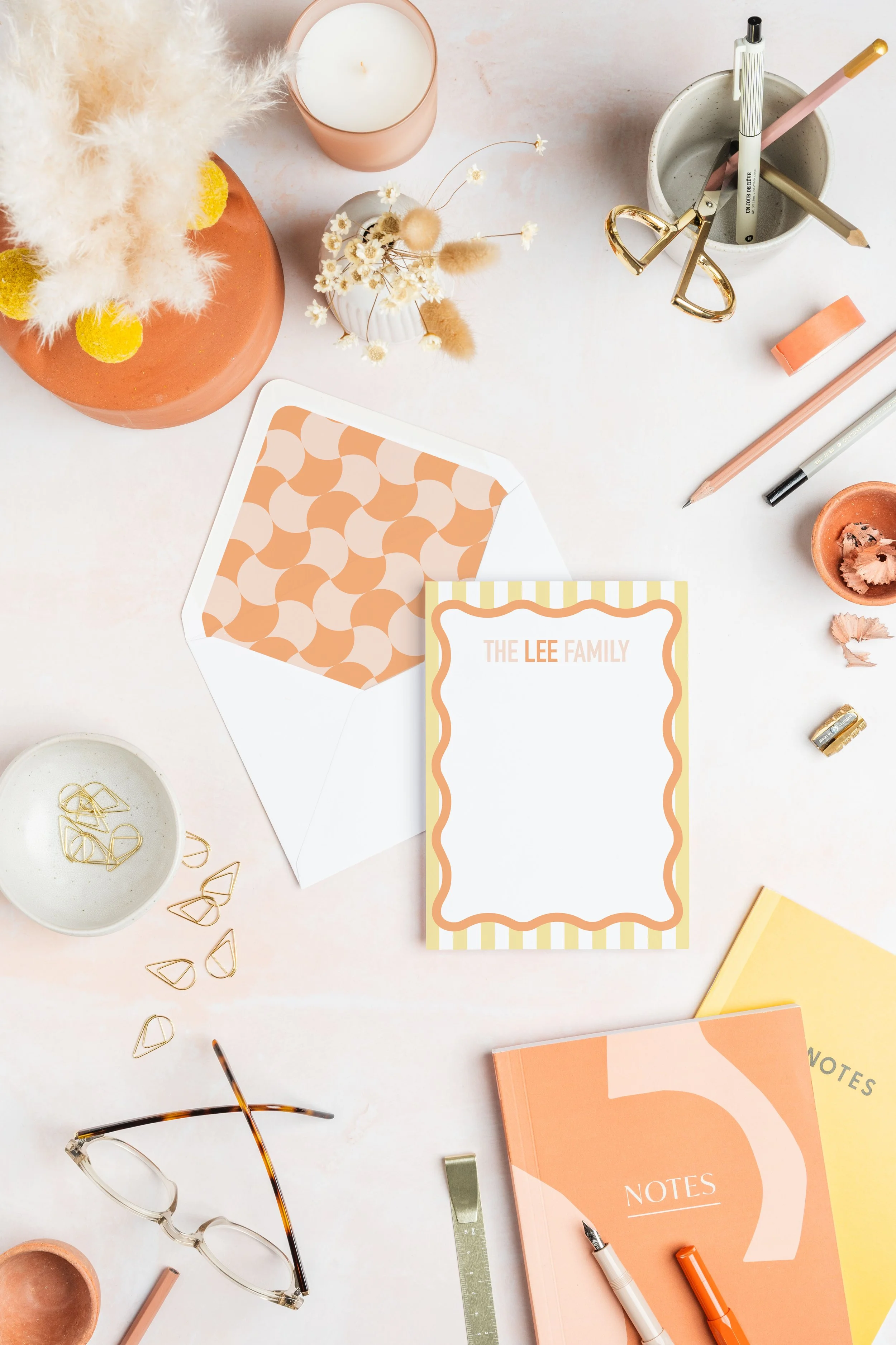 A desk with various stationery items including a peach-colored vase with pampas grass, a small vase with dried flowers, a candle in a pink holder, a pair of gold scissors, pencils and pens in a gray concrete holder, a roll of pink tape, a yellow notepad, and a peach and pink notebook, along with a pair of reading glasses, a ruler, and some loose paper clips.