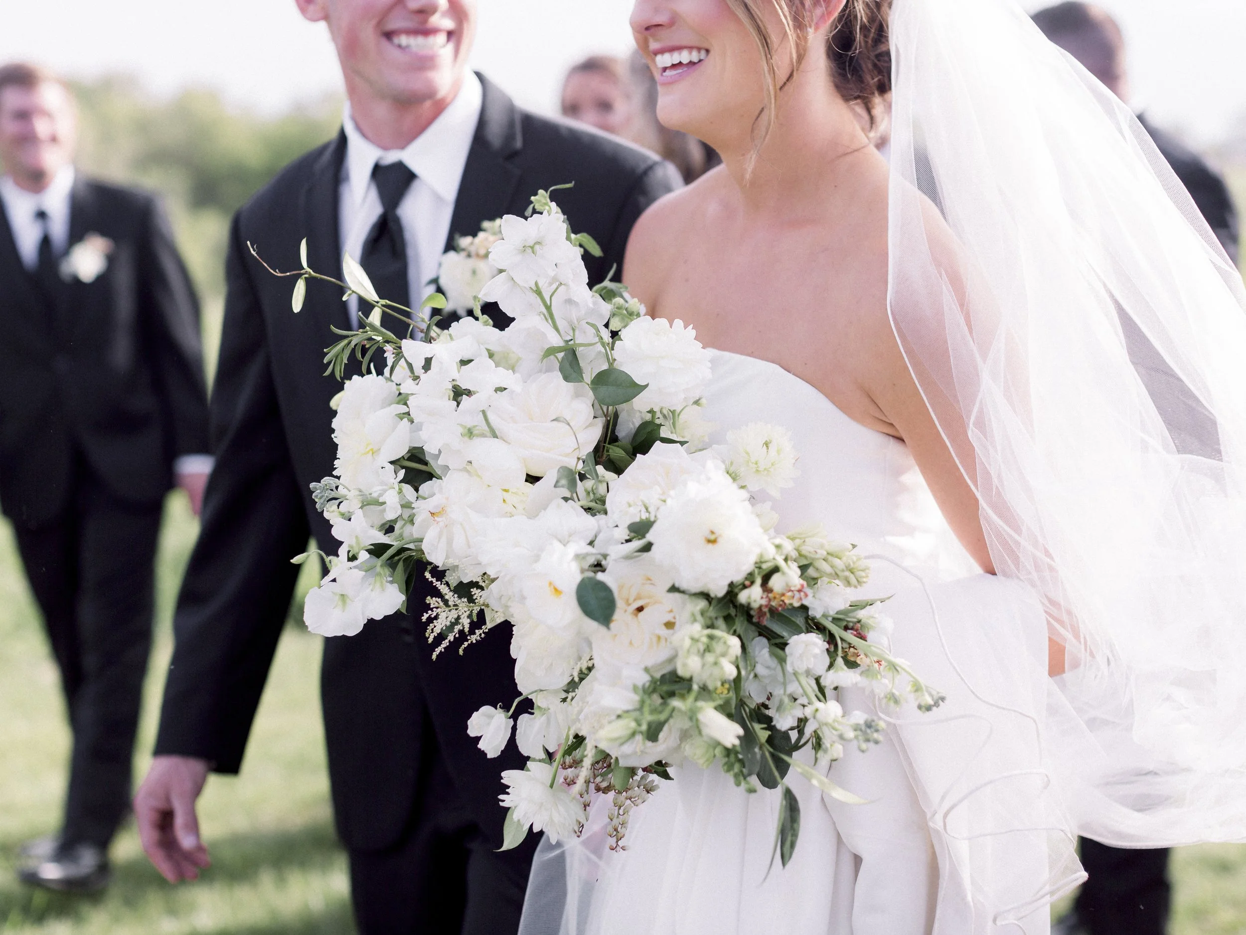 A bride wearing a white wedding gown and veil holding a large bouquet of white flowers, smiling during her wedding ceremony outside. The groom in a black suit with a white shirt and black tie is standing beside her, smiling. Several guests in formal 