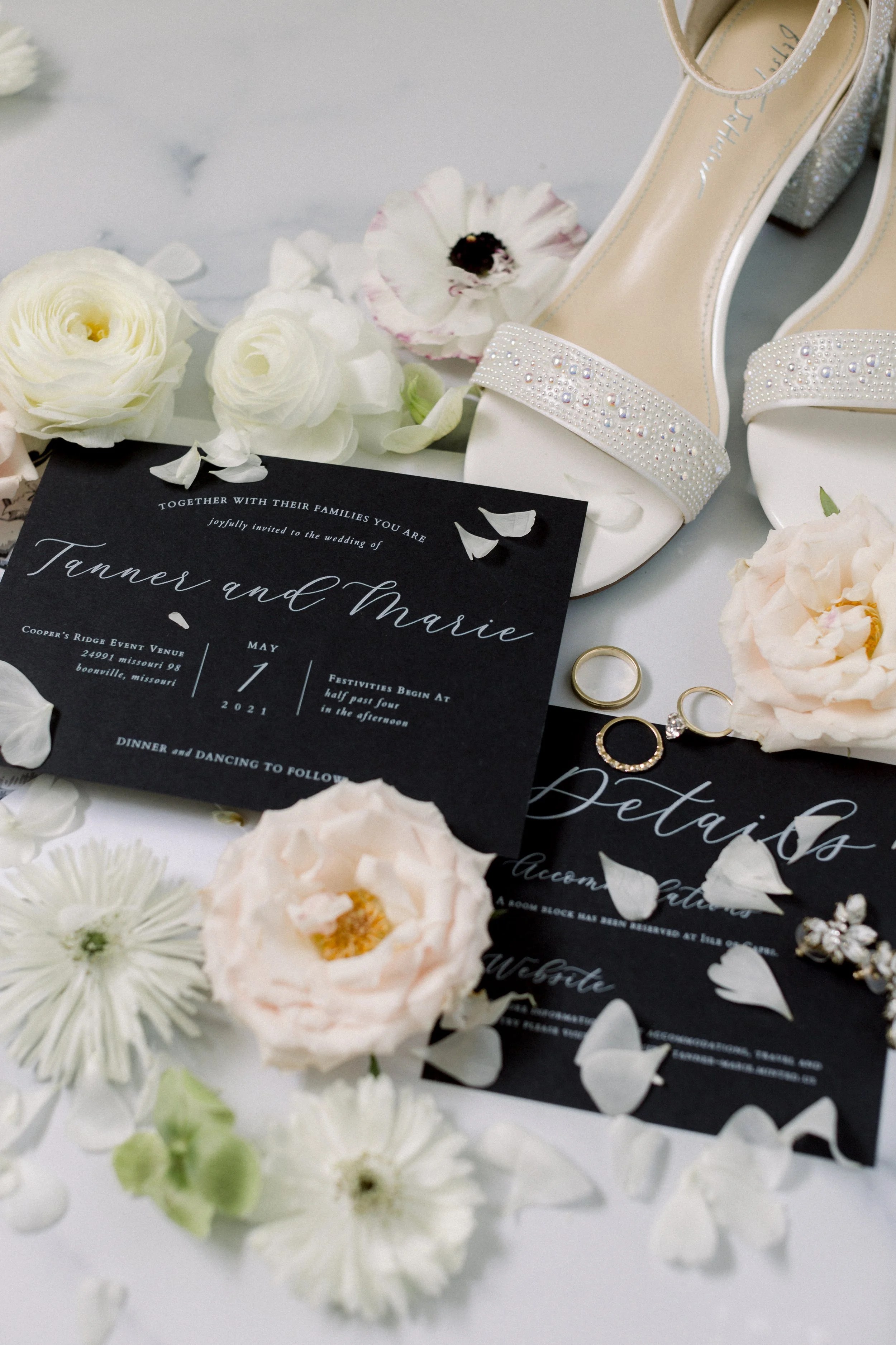 Wedding invitation surrounded by white and light pink flowers, with a pair of white pearl-embellished high-heeled shoes and gold wedding rings placed nearby.