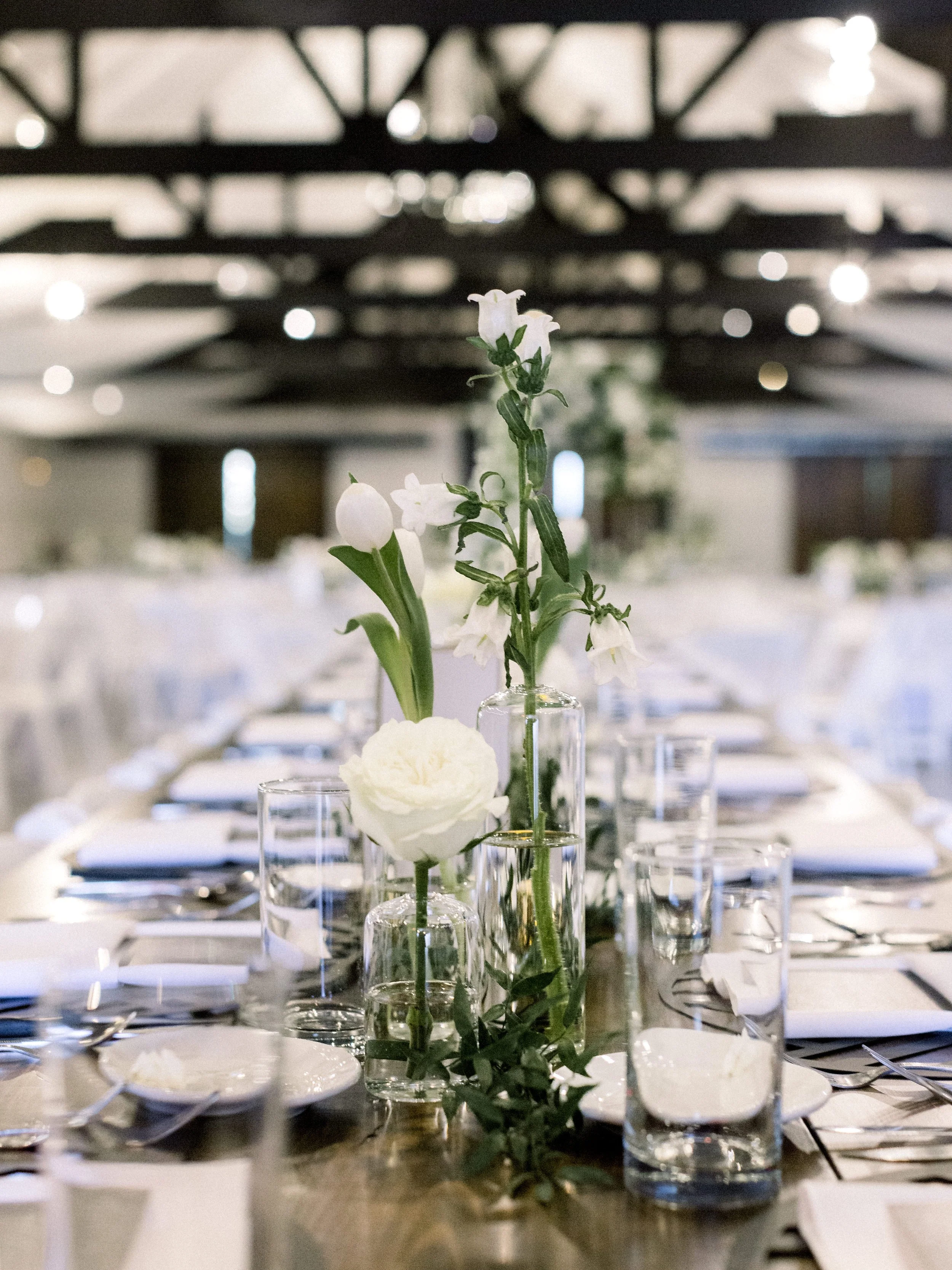 A dining table set with white plates, glasses, silverware, and a floral centerpiece in glass vases, under a dark ceiling with decorative beams.