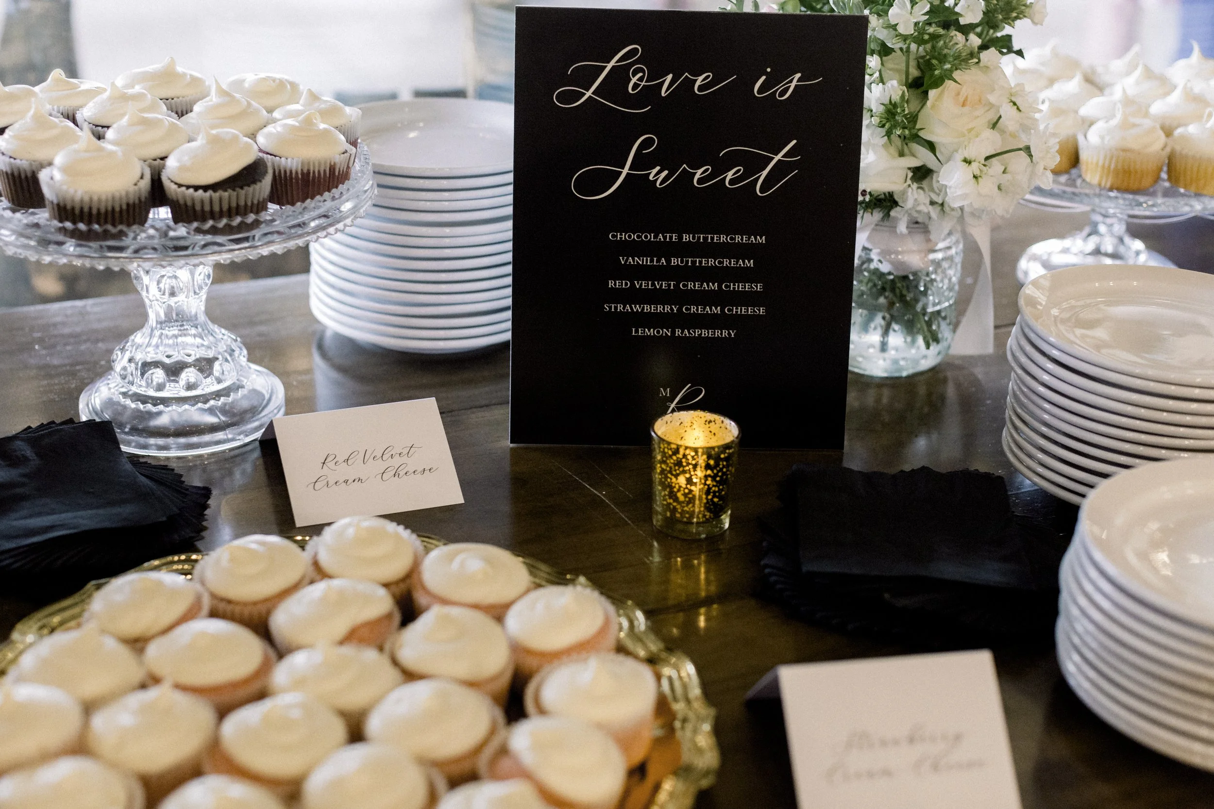 Assorted cupcakes on glass cake stands and plates, with a sign that reads 'Love is sweet' and a guest sign in the background.