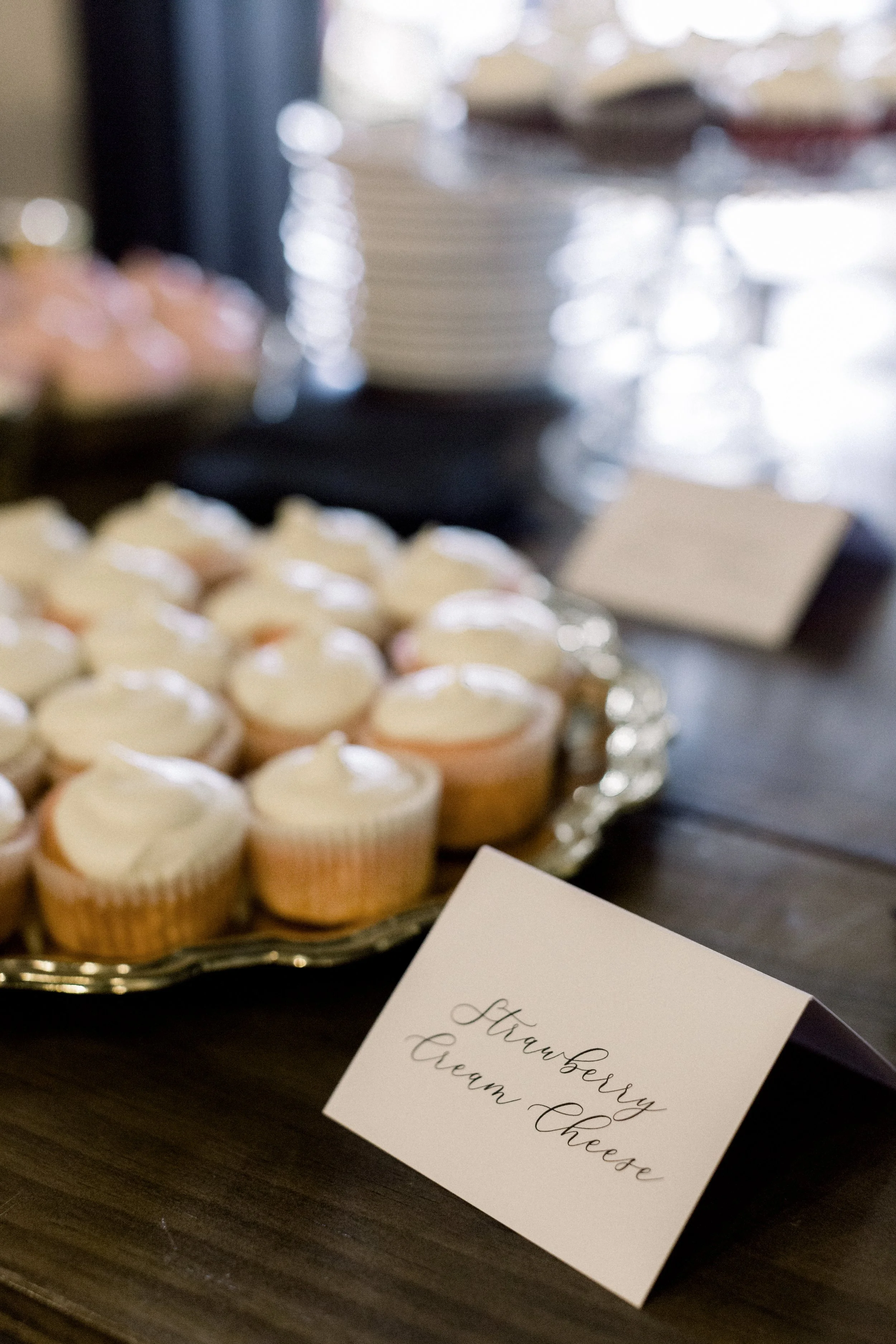 Tray of cupcakes with white frosting on a table, with a blurred background and a card reading "Strawberry Cream Cheese" in the foreground.