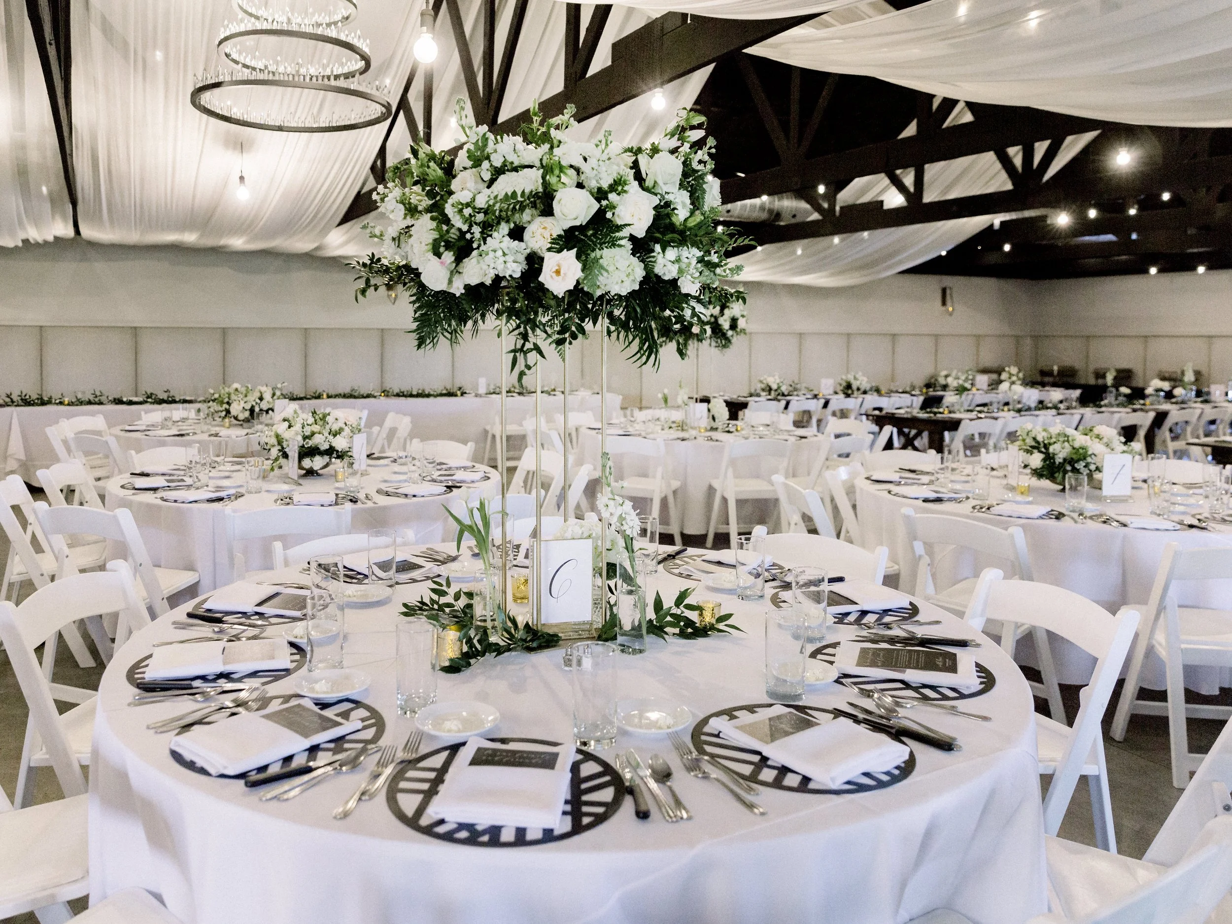 Wedding banquet hall decorated with white tablecloths, floral centerpieces, and white chairs; ceiling draped with white fabric and hanging lights.