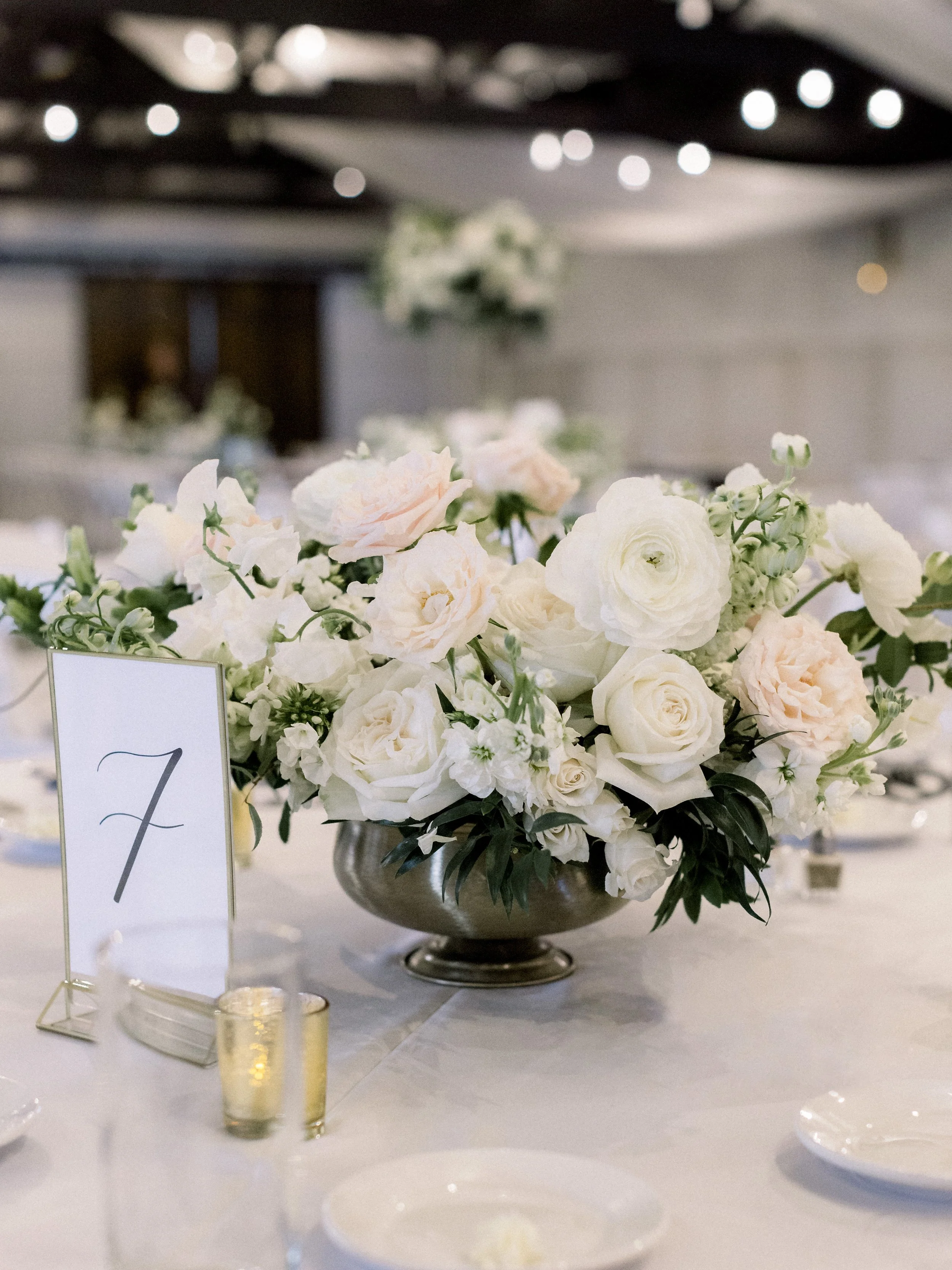 A large floral centerpiece with white and light pink roses and greenery on a table at a wedding or event.