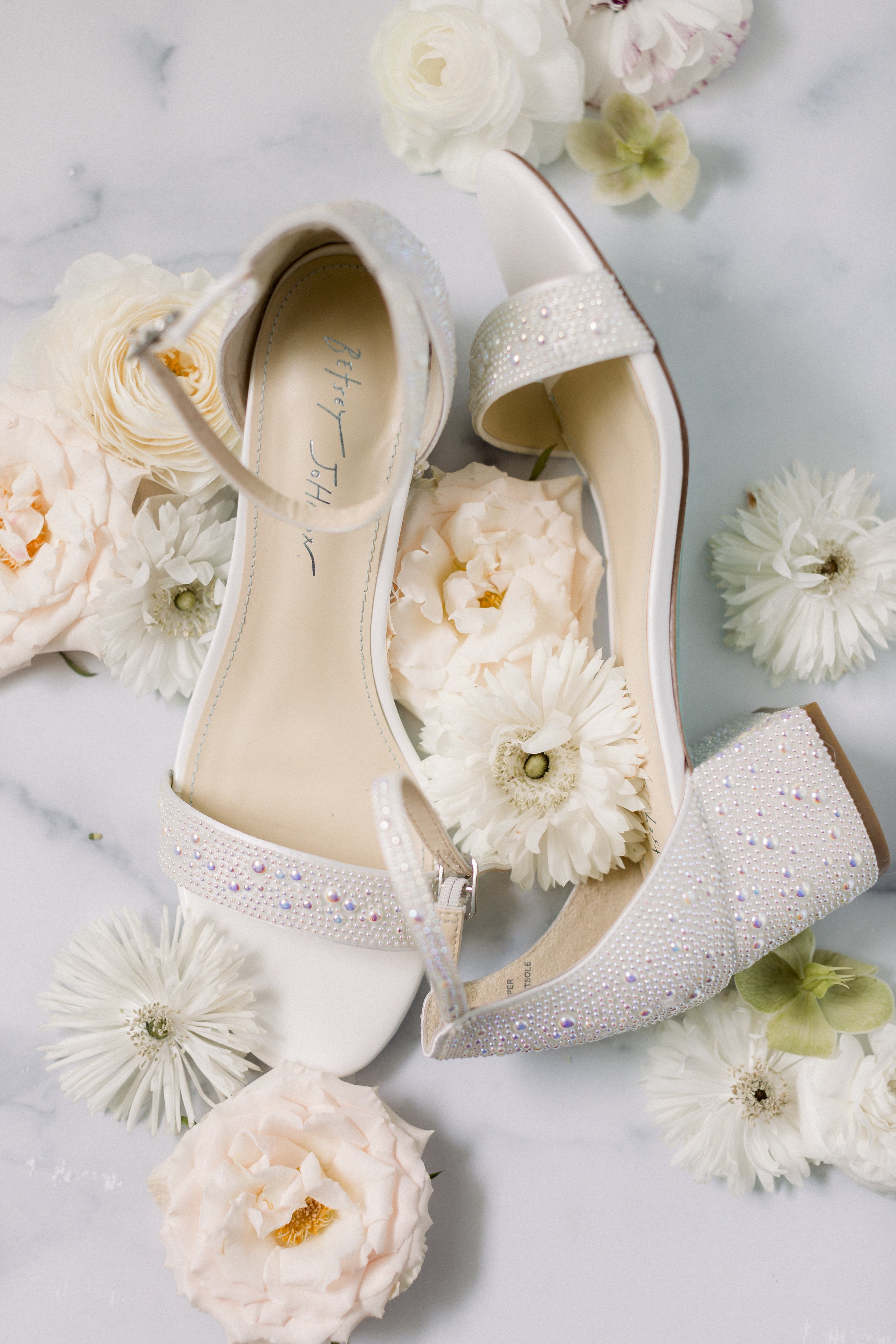 A pair of white bridal shoes with rhinestone embellishments, surrounded by light-colored flowers on a white surface.