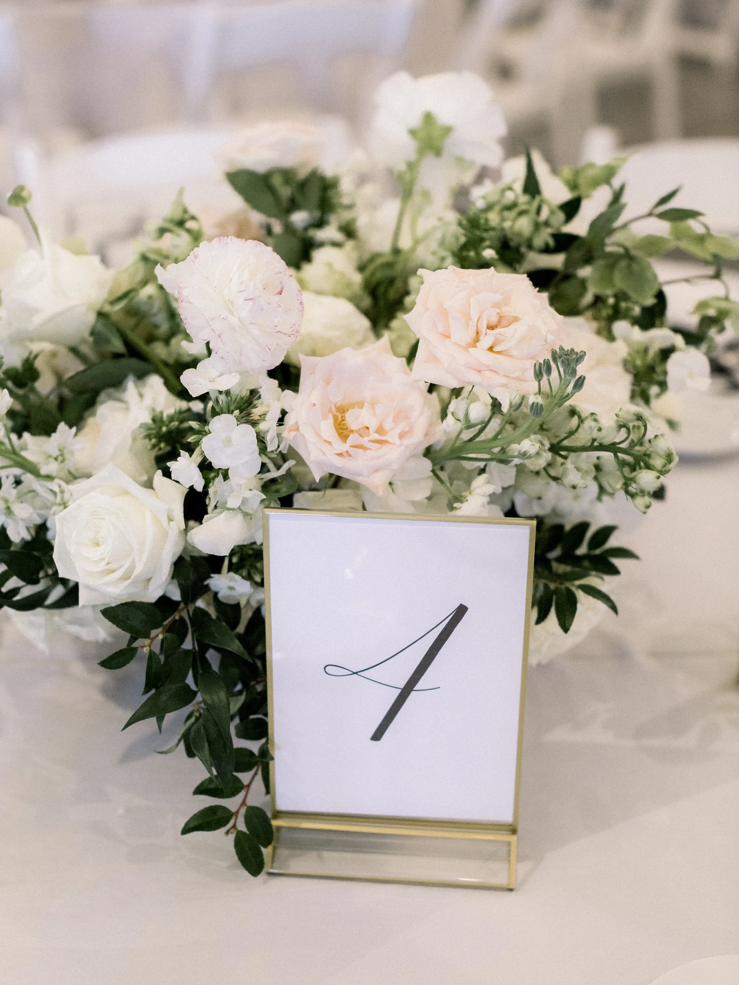 A floral centerpiece with white and soft pink roses, white ranunculus, and greenery, with a table number card displaying the number 4, on a white table at a wedding or event.