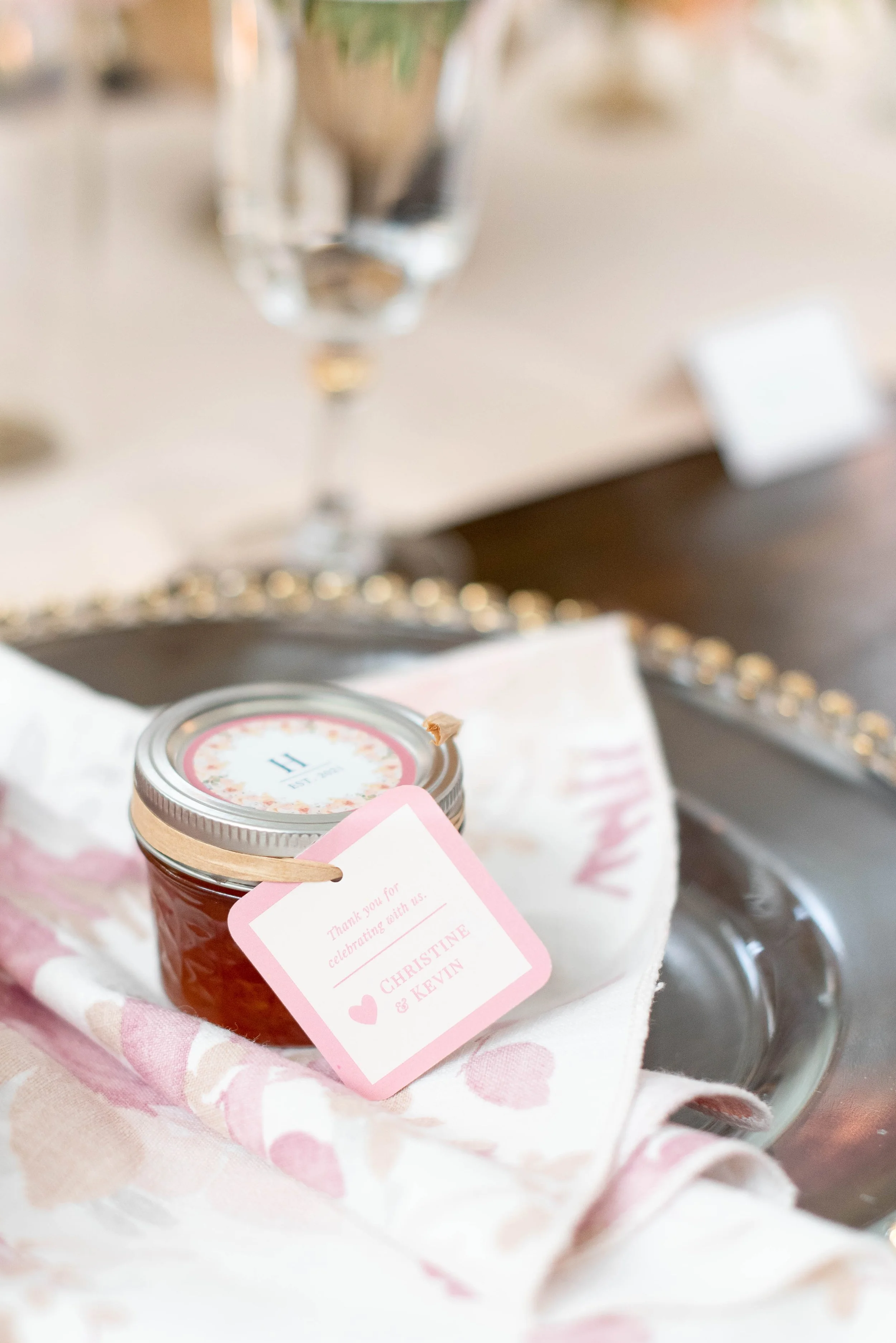 A small jar of jam with a pink tag, set on a pink cloth with heart patterns, surrounded by gift bags and a glass vase in the background.