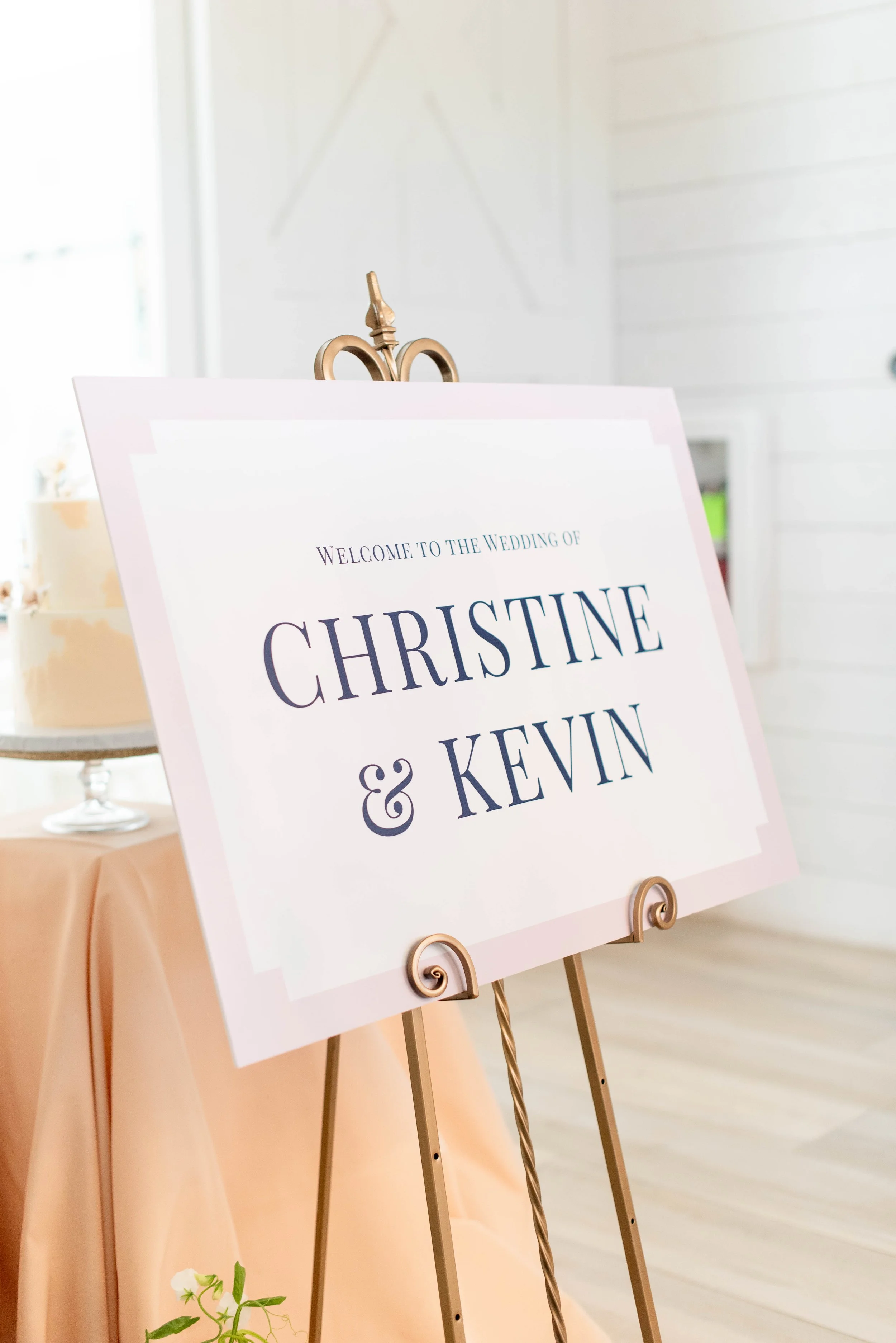 Wedding welcome sign with names Christine & Kevin on a gold easel, placed on a table with peach-colored tablecloth in a bright room.
