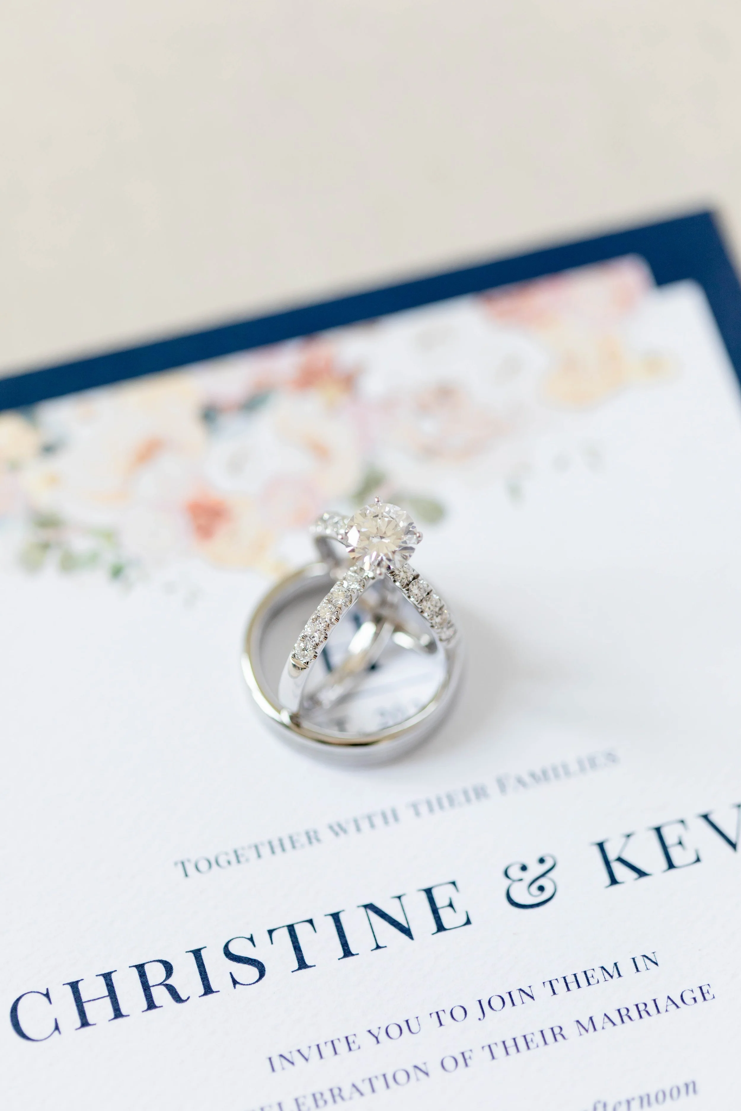 Close-up of wedding rings placed on a wedding invitation with floral decorations in the background.