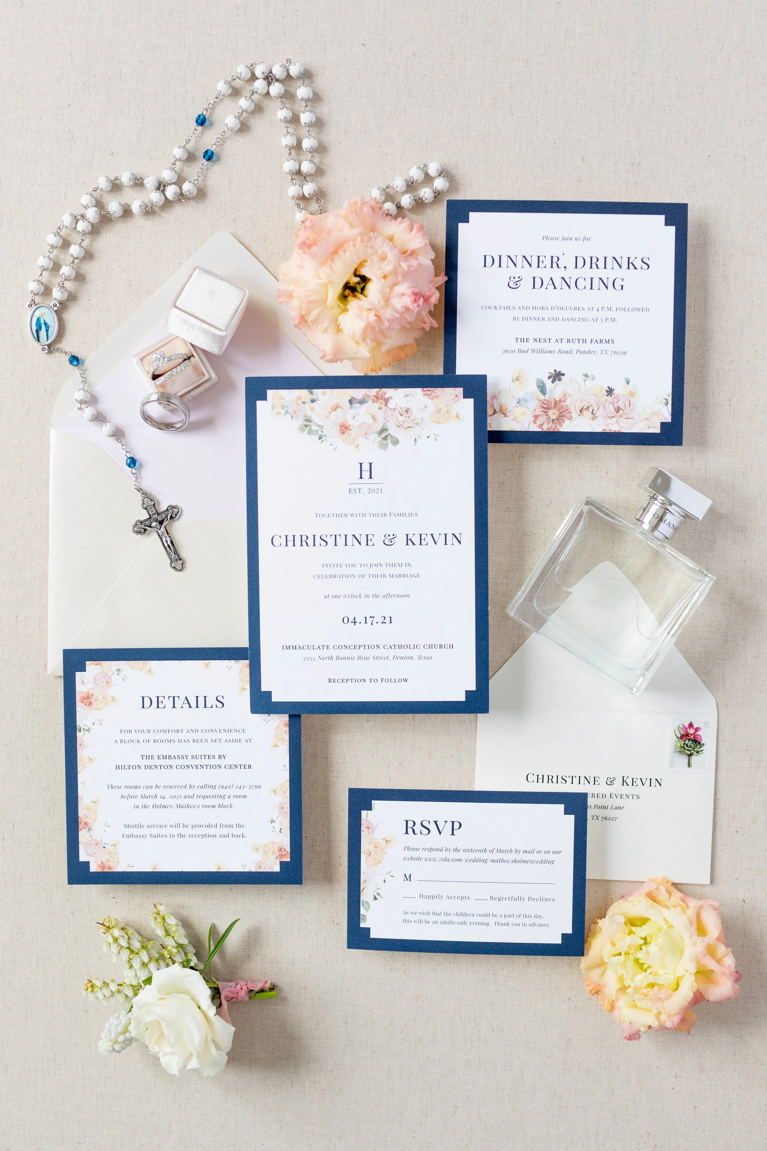 Wedding invitation suite with navy blue borders, floral accents, and event details, surrounded by pink and cream flowers, a rosary, rings in a white box, a perfume bottle, and small floral boutonniere.