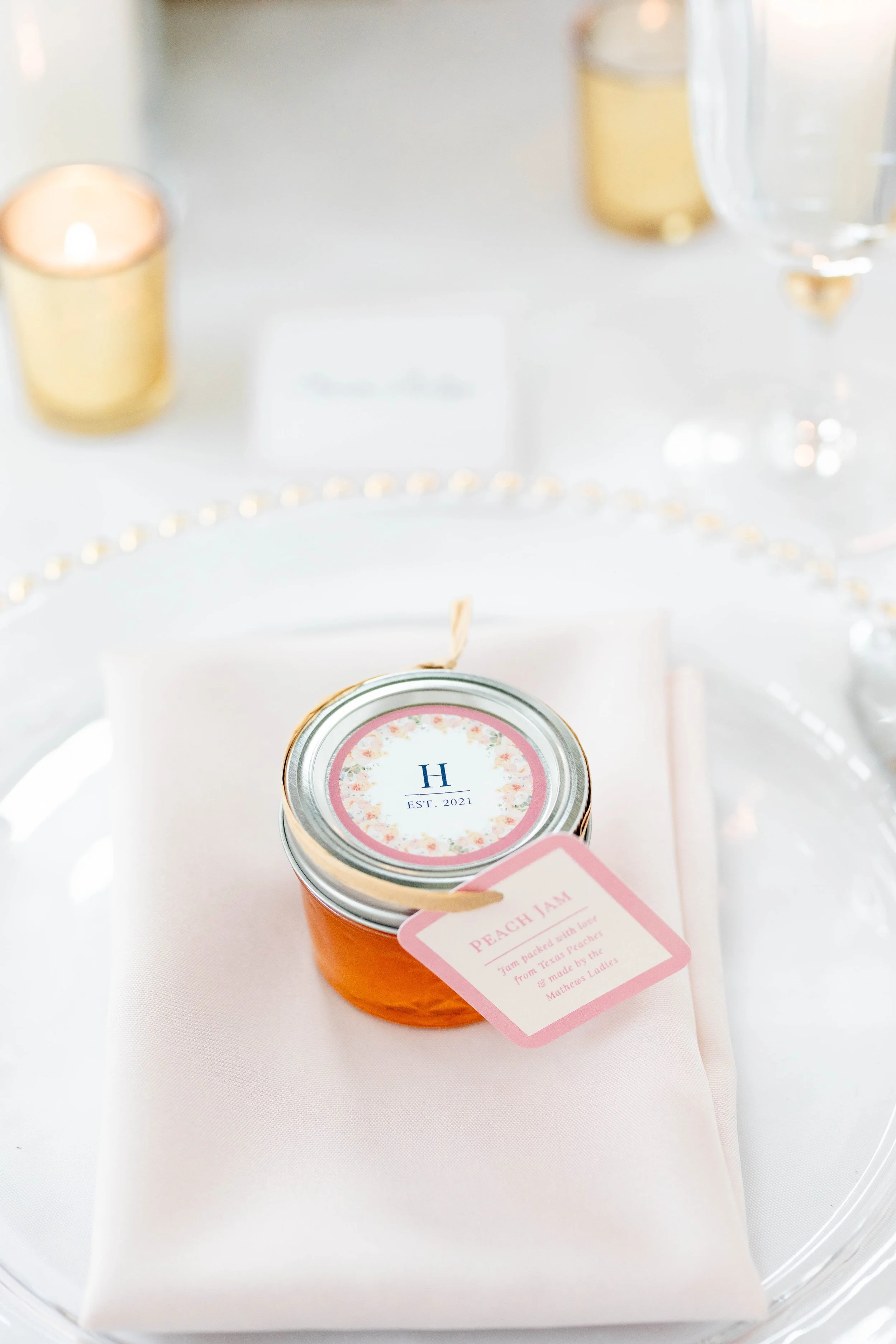 A small orange jar of peach jam with a pink label hanging from its lid, on a white surface, with a white background and some gold decorations and candles in the background.