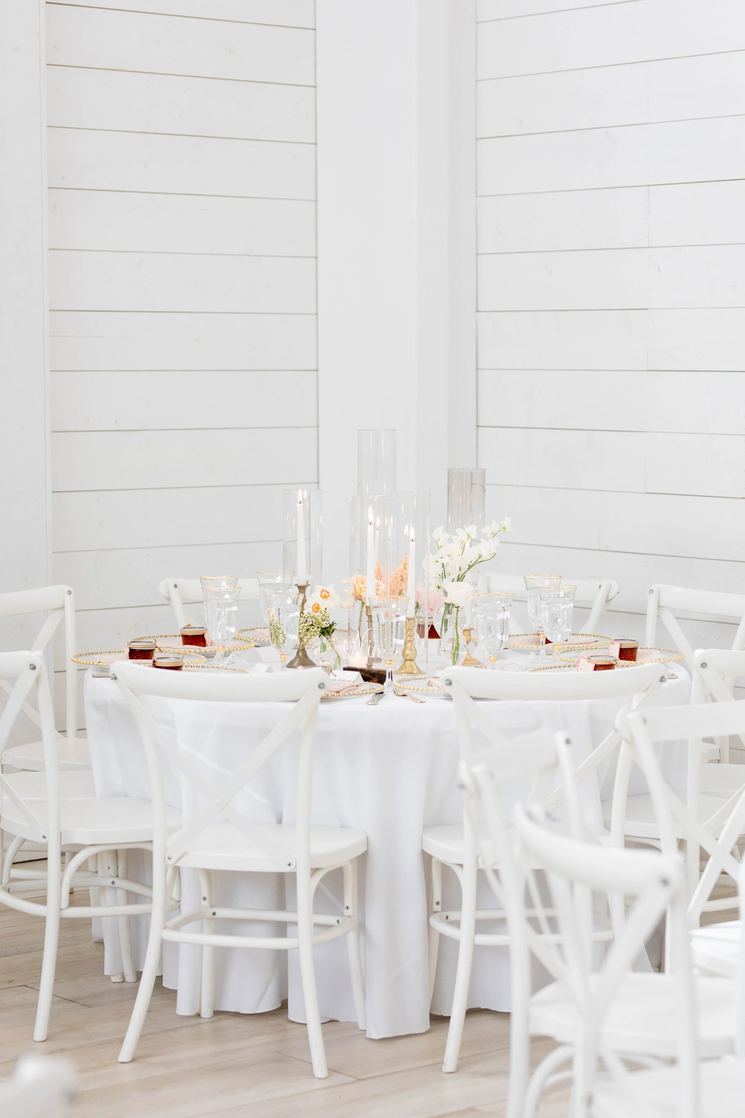 Elegant event setting with round table covered in white tablecloth, surrounded by white chairs, featuring floral arrangements, candles, and glassware in a bright, white room with shiplap walls.