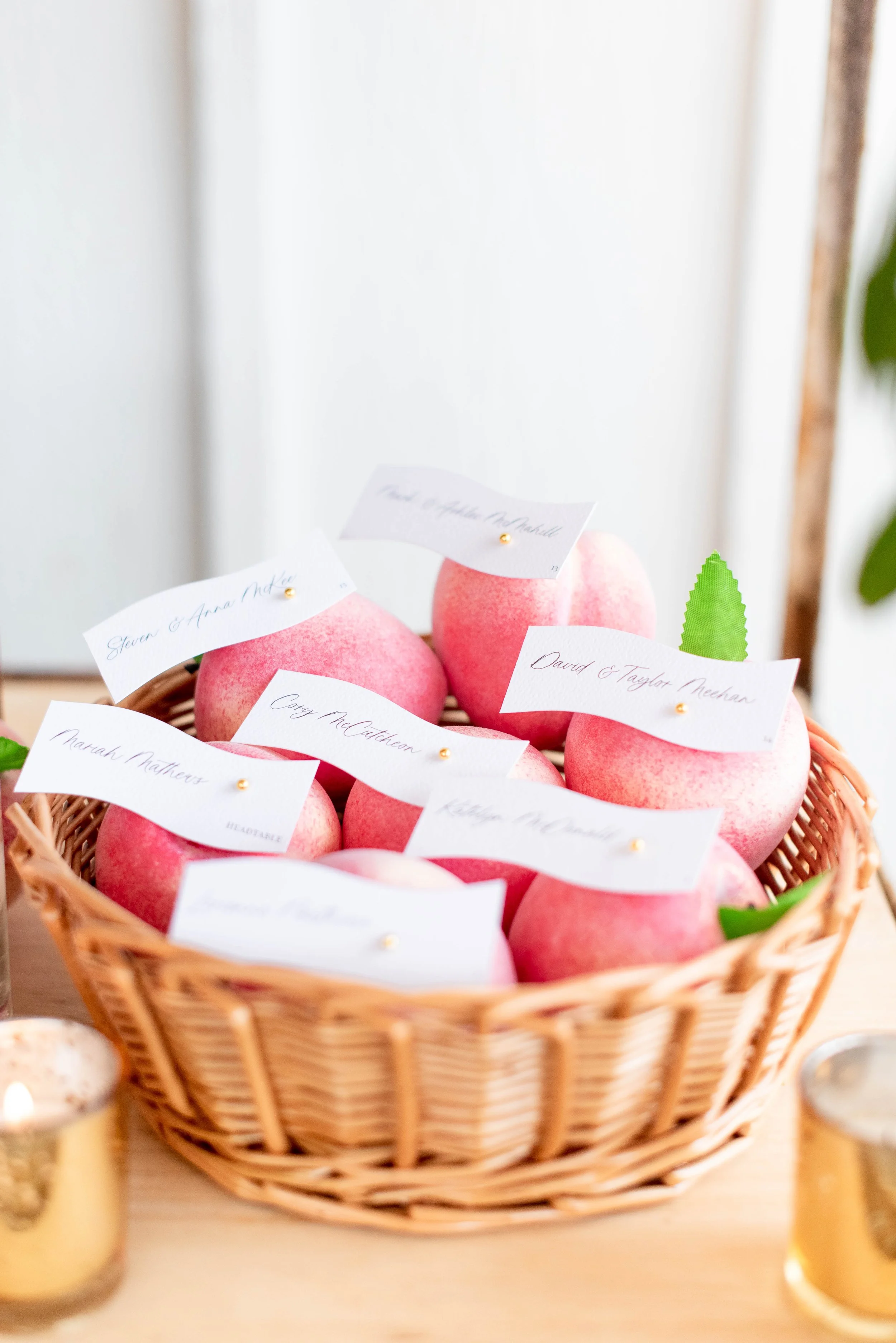 Basket of pink apples with small notes attached, on a light-colored table, with candles on either side.