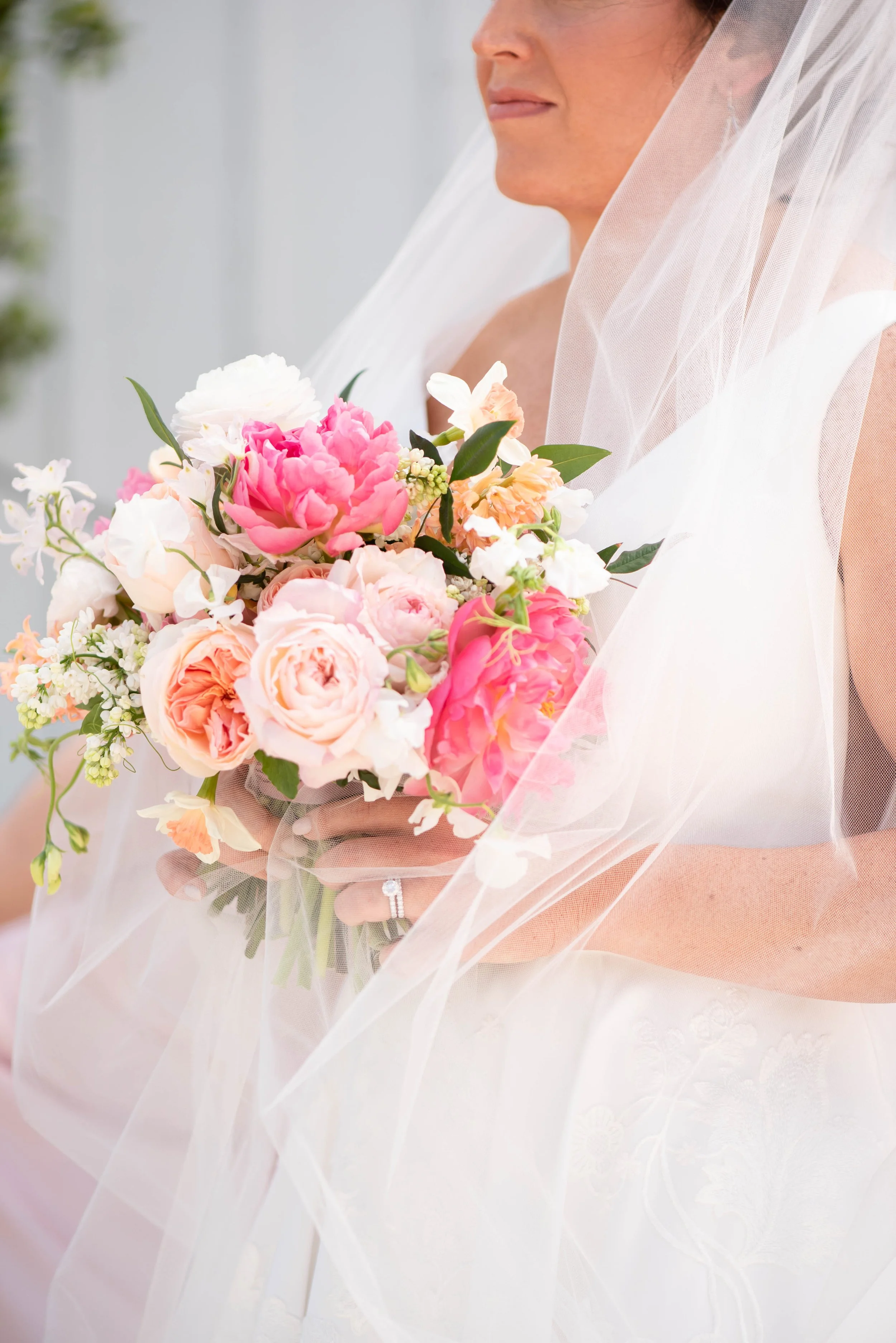 Close-up of a bride holding a colorful bouquet of pink, white, and peach flowers, with a sheer white veil over her shoulders. The bride is smiling softly, with a light-colored dress and a wedding ring visible on her finger.