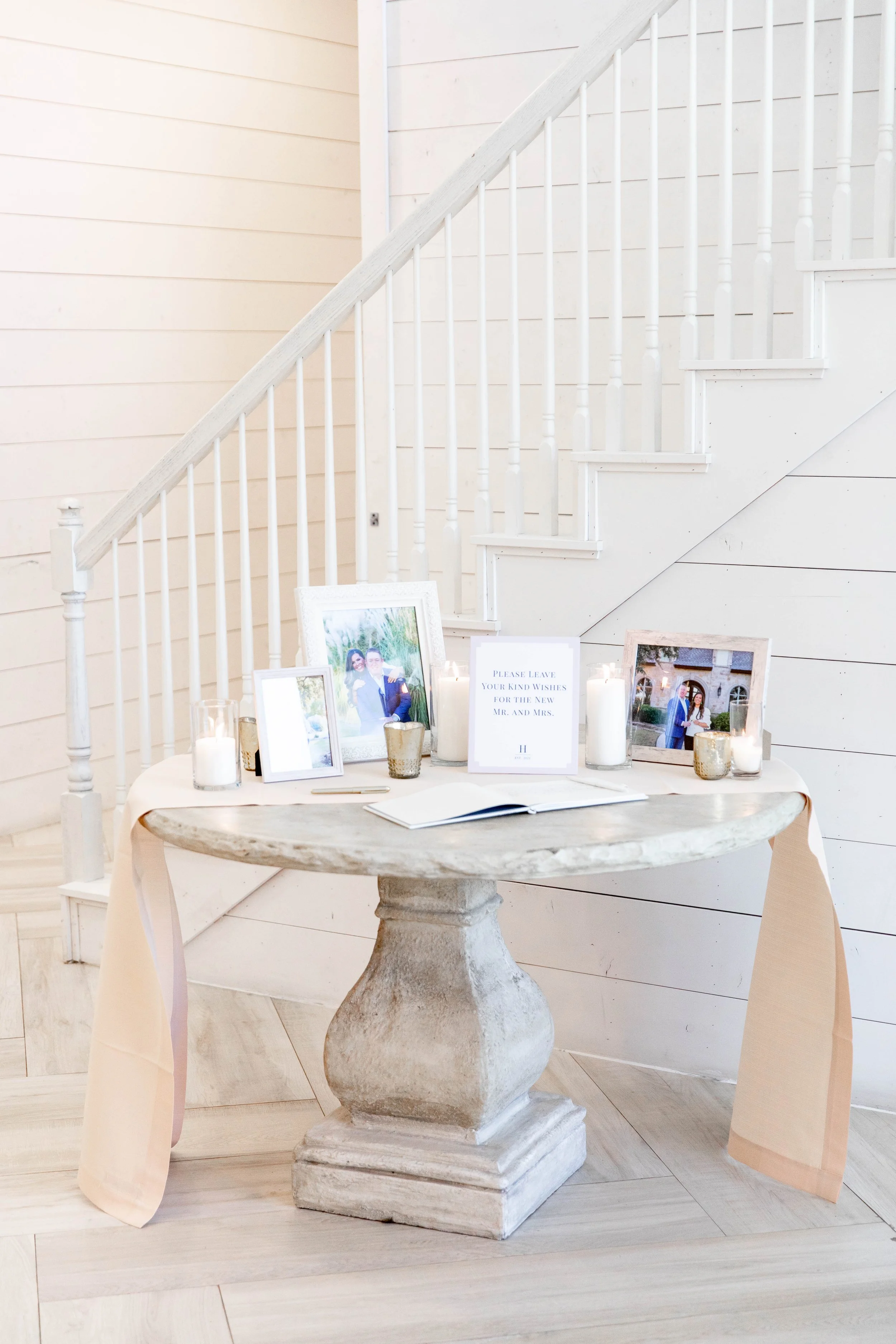 A round stone table with a stack of framed wedding photos, white candles, and a sign for guests, positioned in front of a white staircase with horizontal wood paneling in the background.