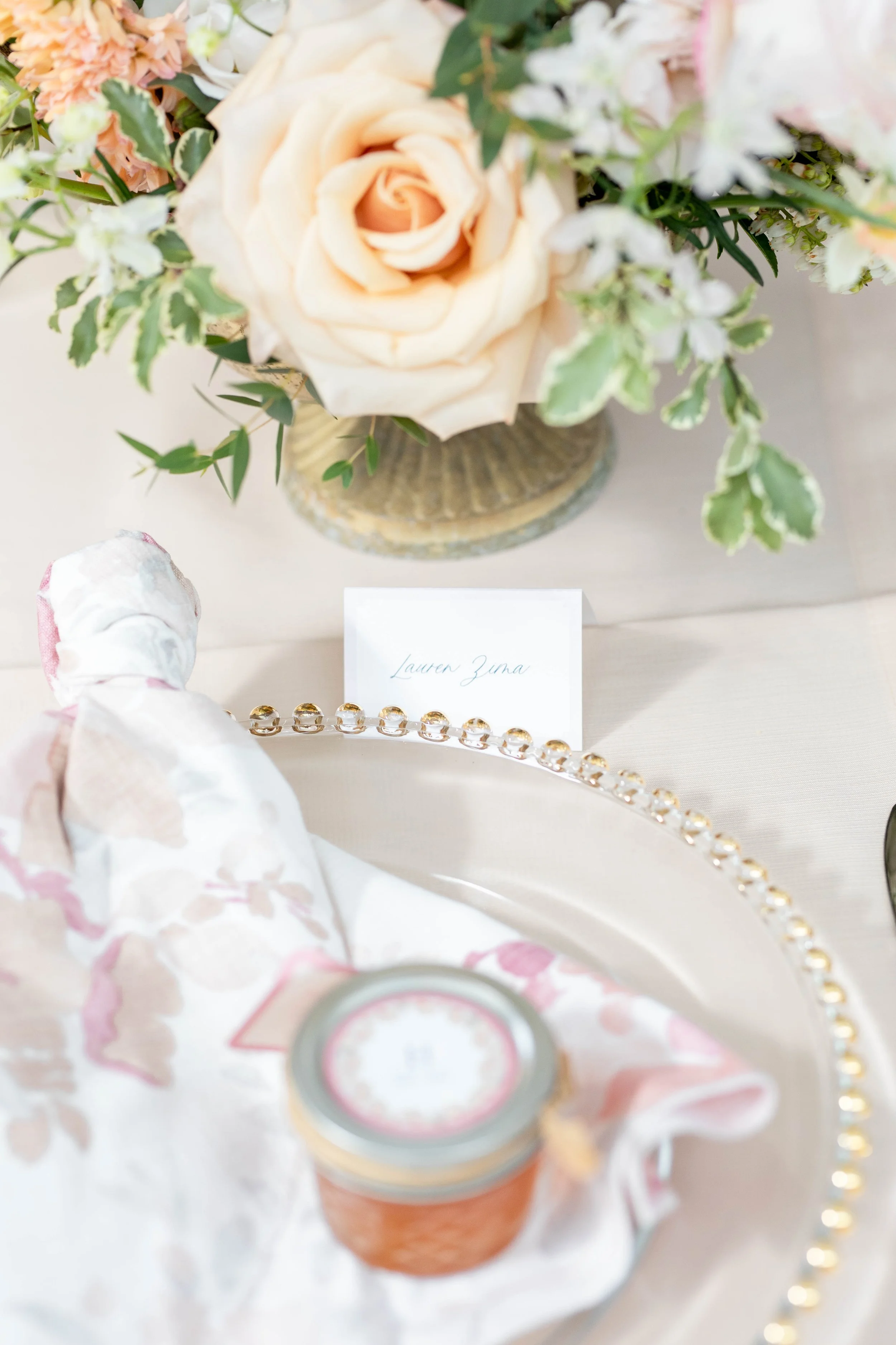 A close-up of a floral arrangement with a peach-colored rose in a vase, surrounded by white and green foliage, and some decorative beads and a small sign visible in the foreground.