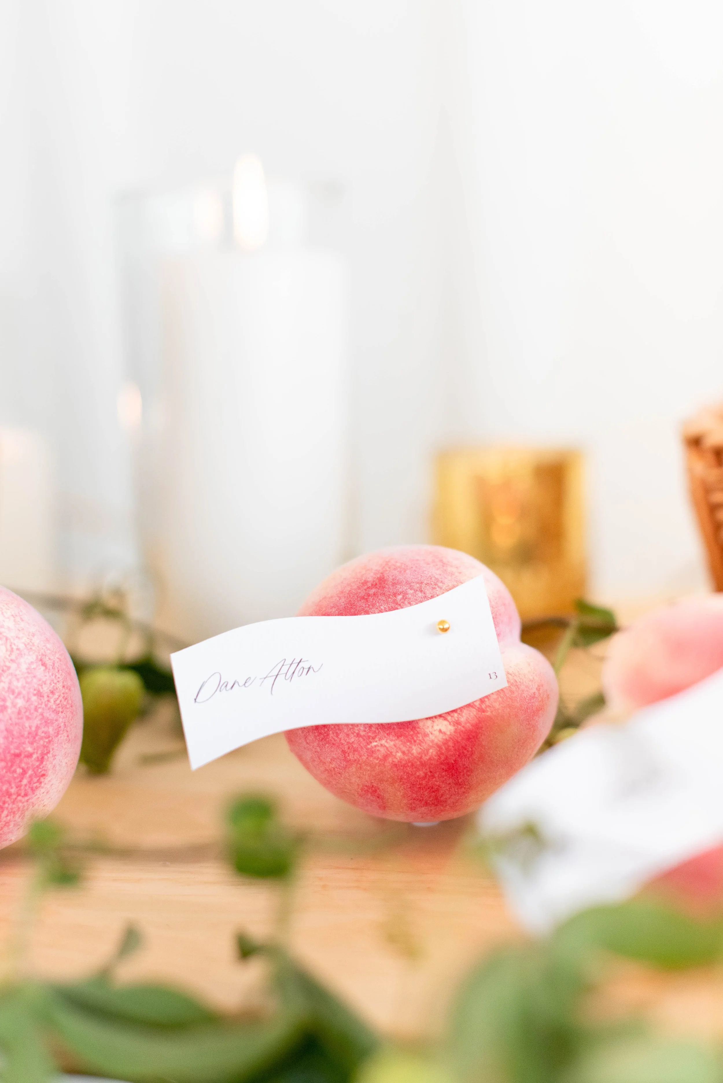 Close-up of pink peaches with a label that says "Dane Alton" on it. Blurred background showing additional peaches and greenery.