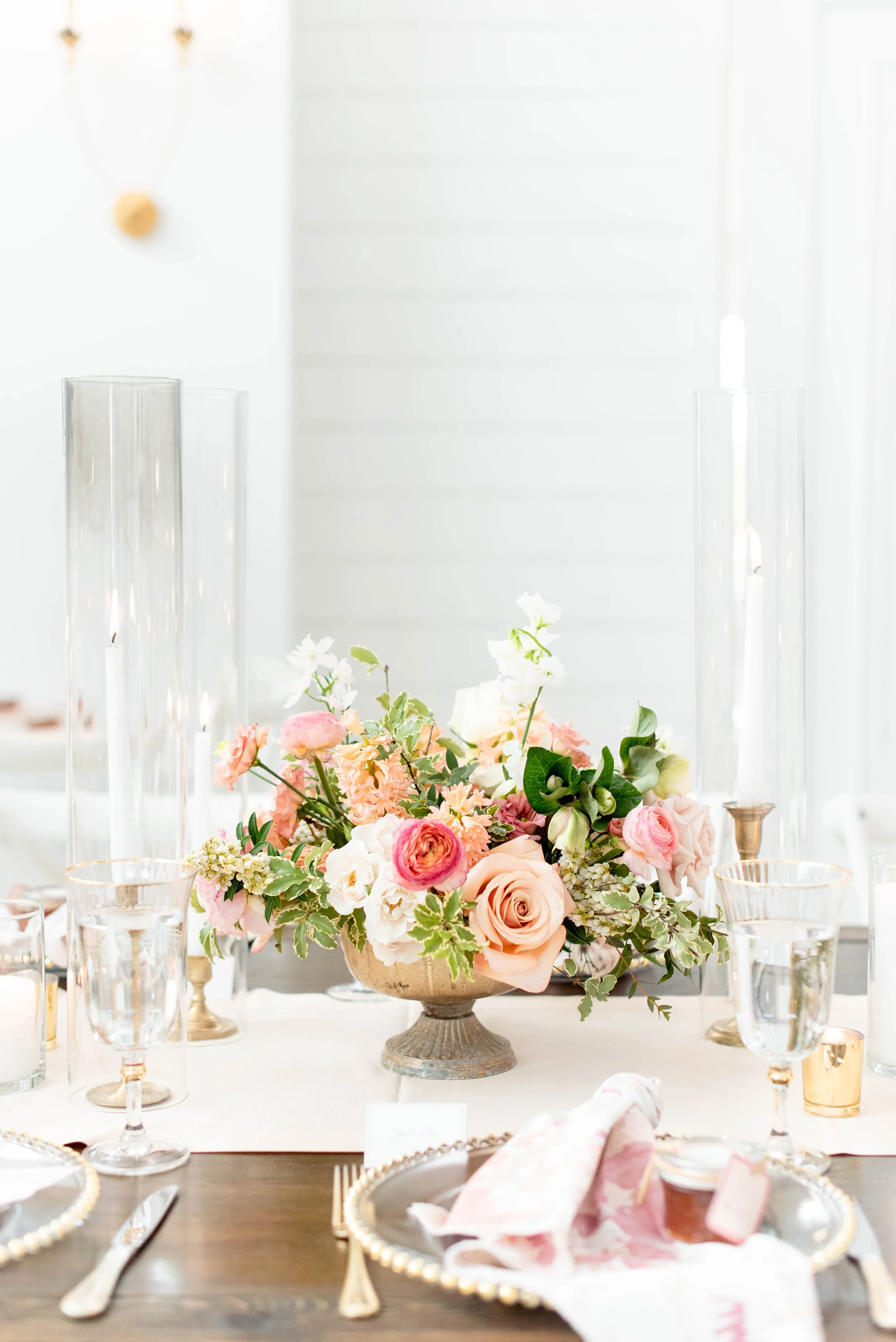 Elegant table setting with a large floral centerpiece featuring pink, white, and peach roses, set on a wooden table with glassware, gold-rimmed chargers, and pink napkins, in a bright, white decorated room.