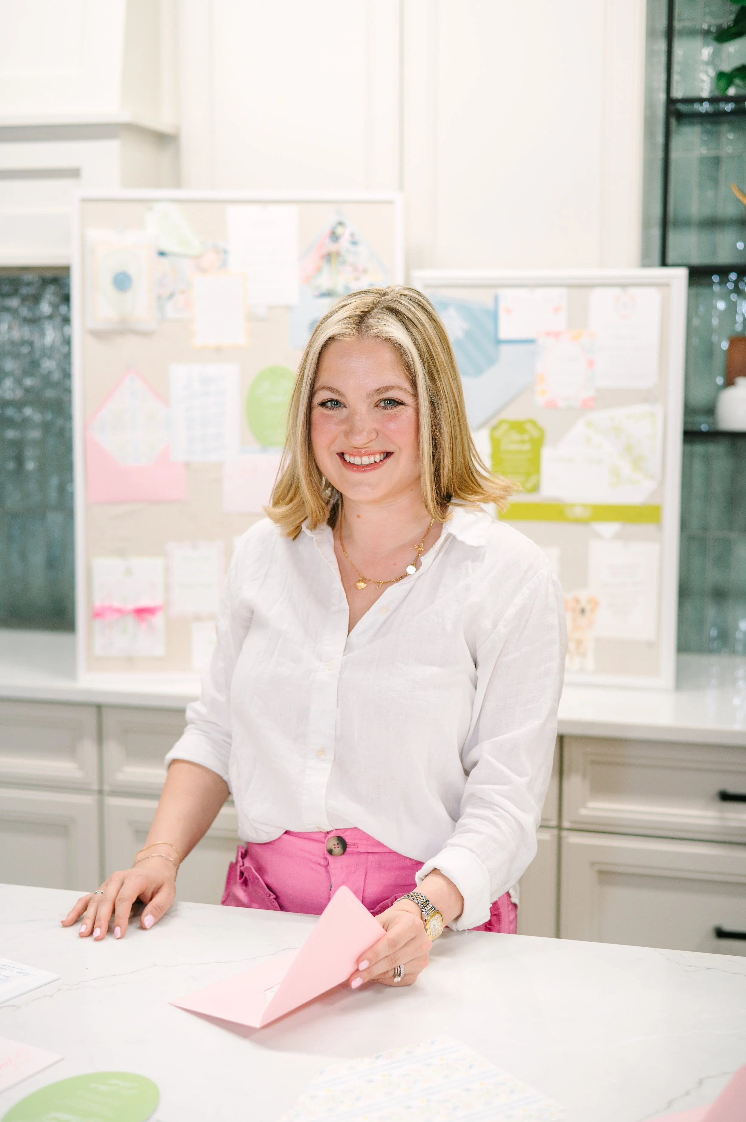 A woman with blonde hair wearing a white blouse and pink pants, smiling and folding pink paper at a table in a bright room with a whiteboard in the background.