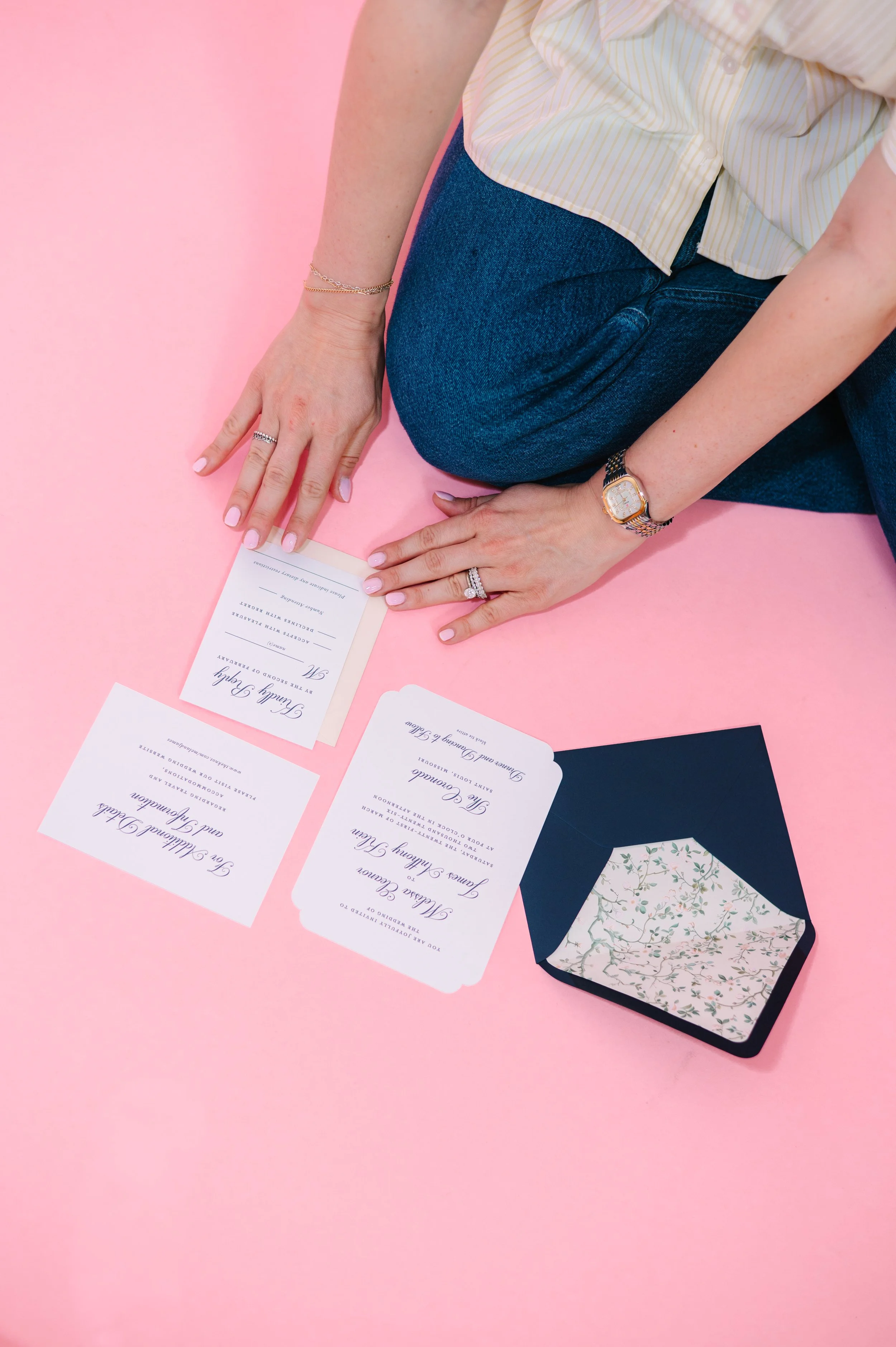 Person kneeling on a pink surface, arranging wedding invitations and wedding stationery, wearing a light yellow striped shirt, dark blue jeans, and a gold watch.