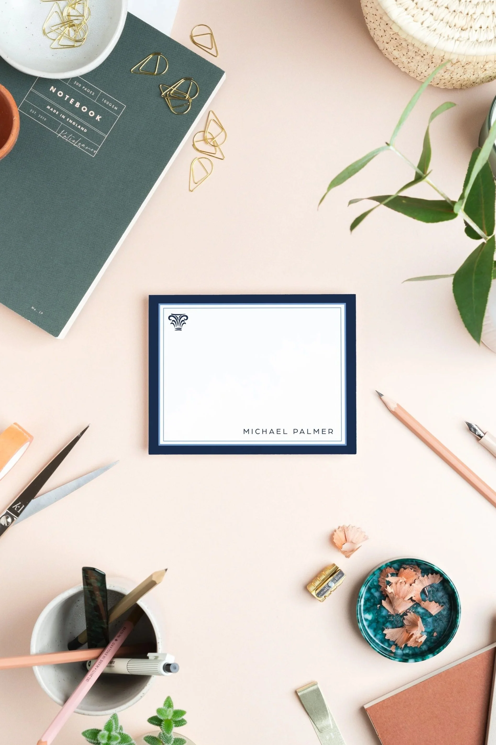 Flat lay of a workspace with a closed blue book titled 'Michael Palmer' on a pink desk, surrounded by stationery, scissors, a notebook, and a small potted plant, with a bowl of pencil shavings and a cup of pens and pencils.