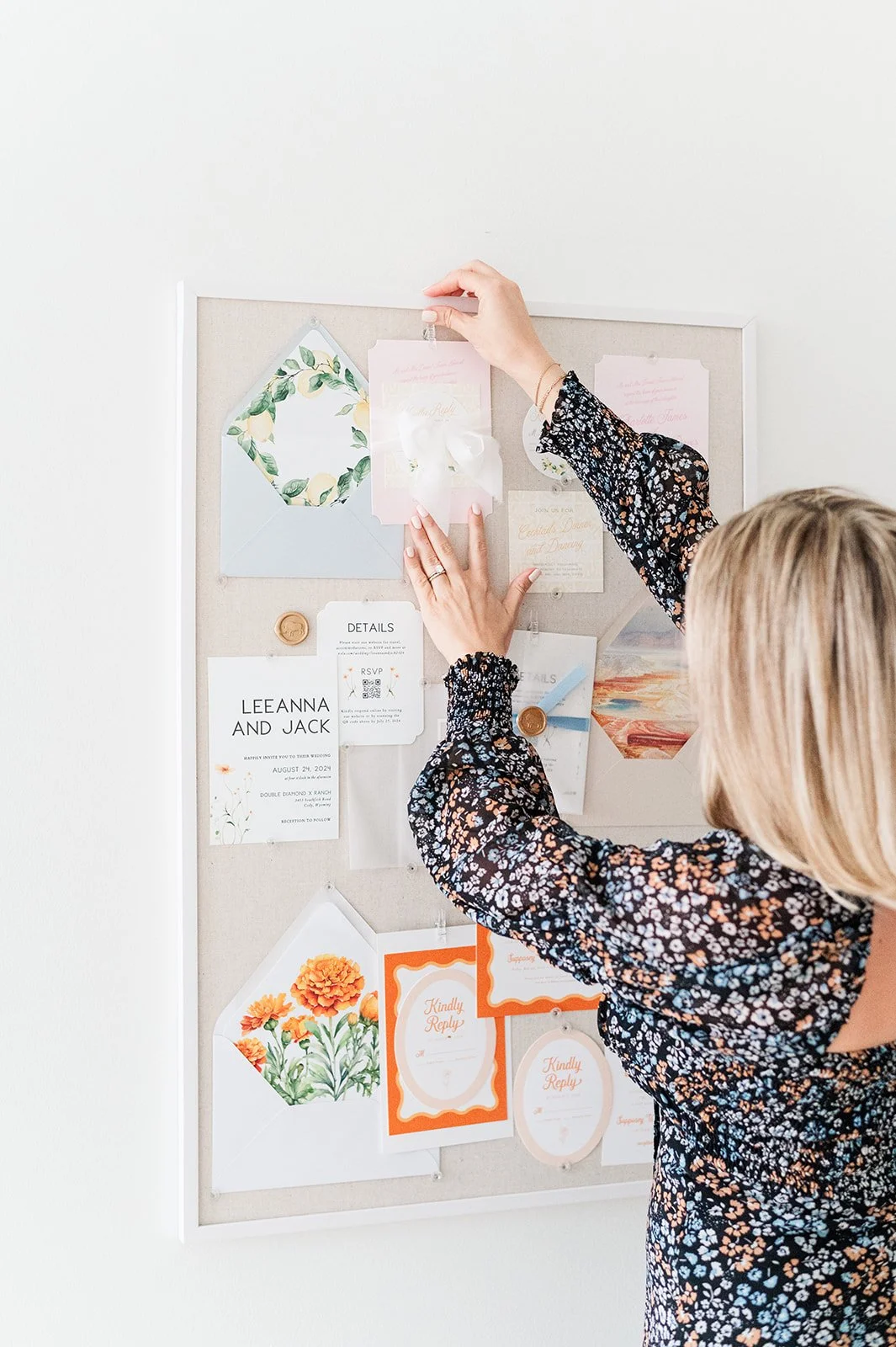 A woman in a black floral dress pinning a pink invitation to a beige wedding or event board, which has various cards, envelopes, and decorative items pinned to it.