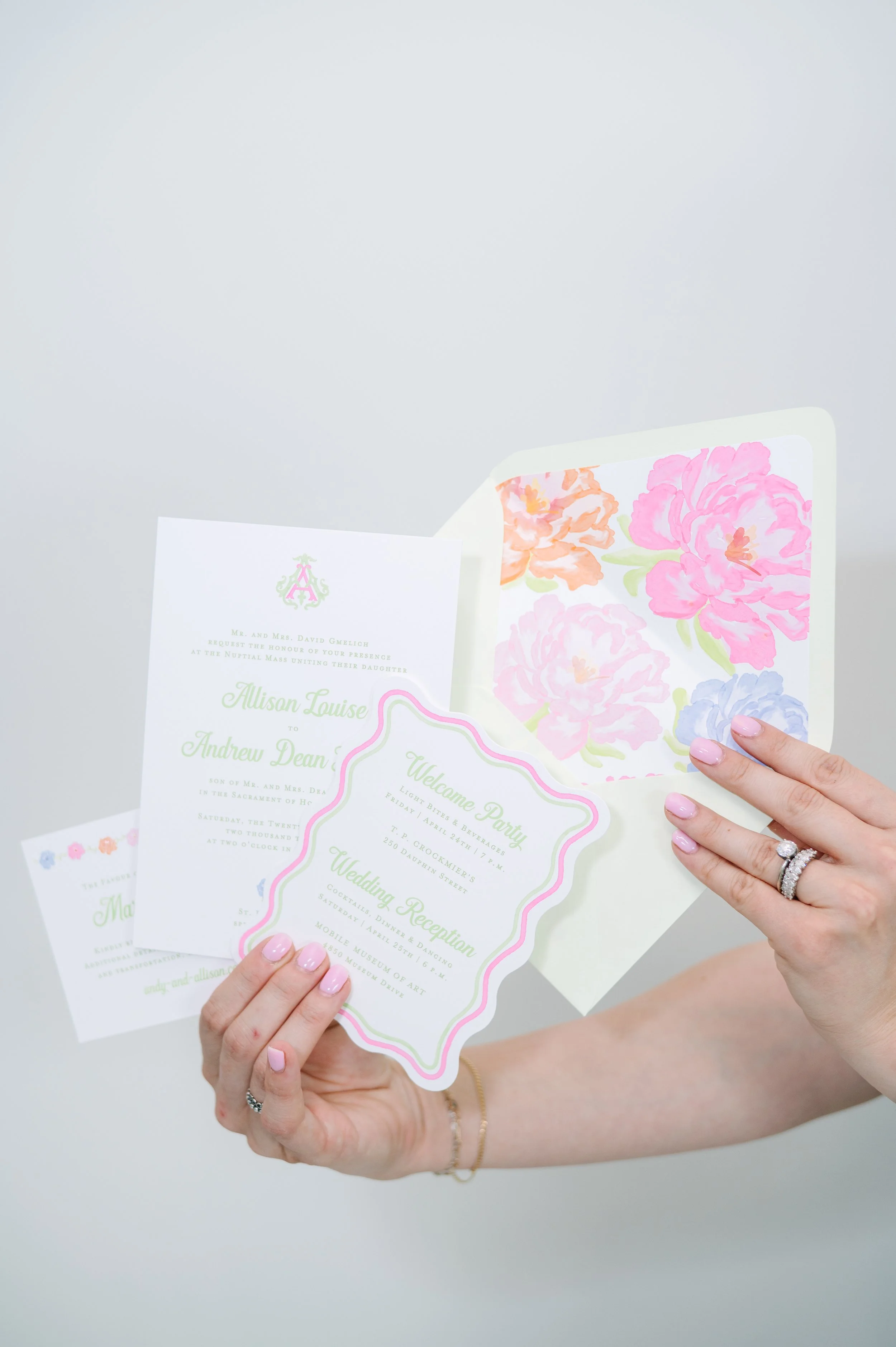A woman holding a wedding invitation with floral artwork and pastel green text, wearing multiple rings and a bracelet.