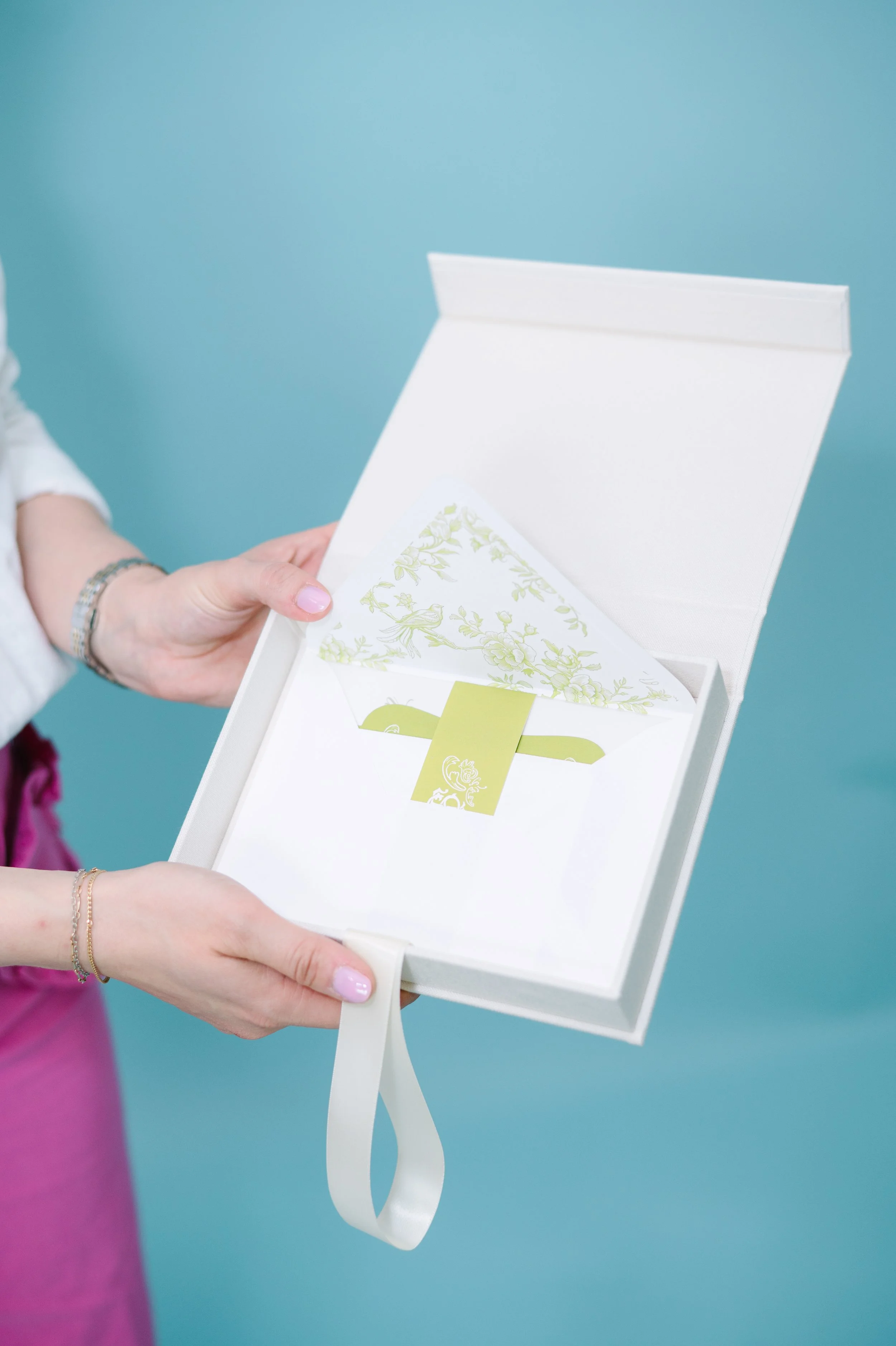 A person holding a white gift box with a floral card inside, against a teal background.