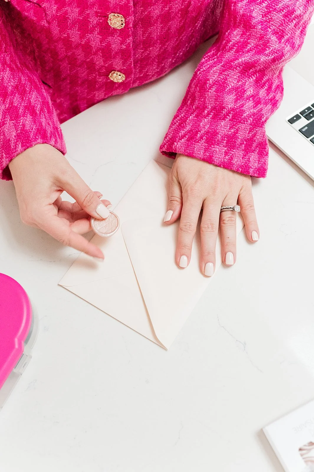 Person in a pink textured blazer holding a wax seal and stamping an envelope on a white table with a laptop nearby.
