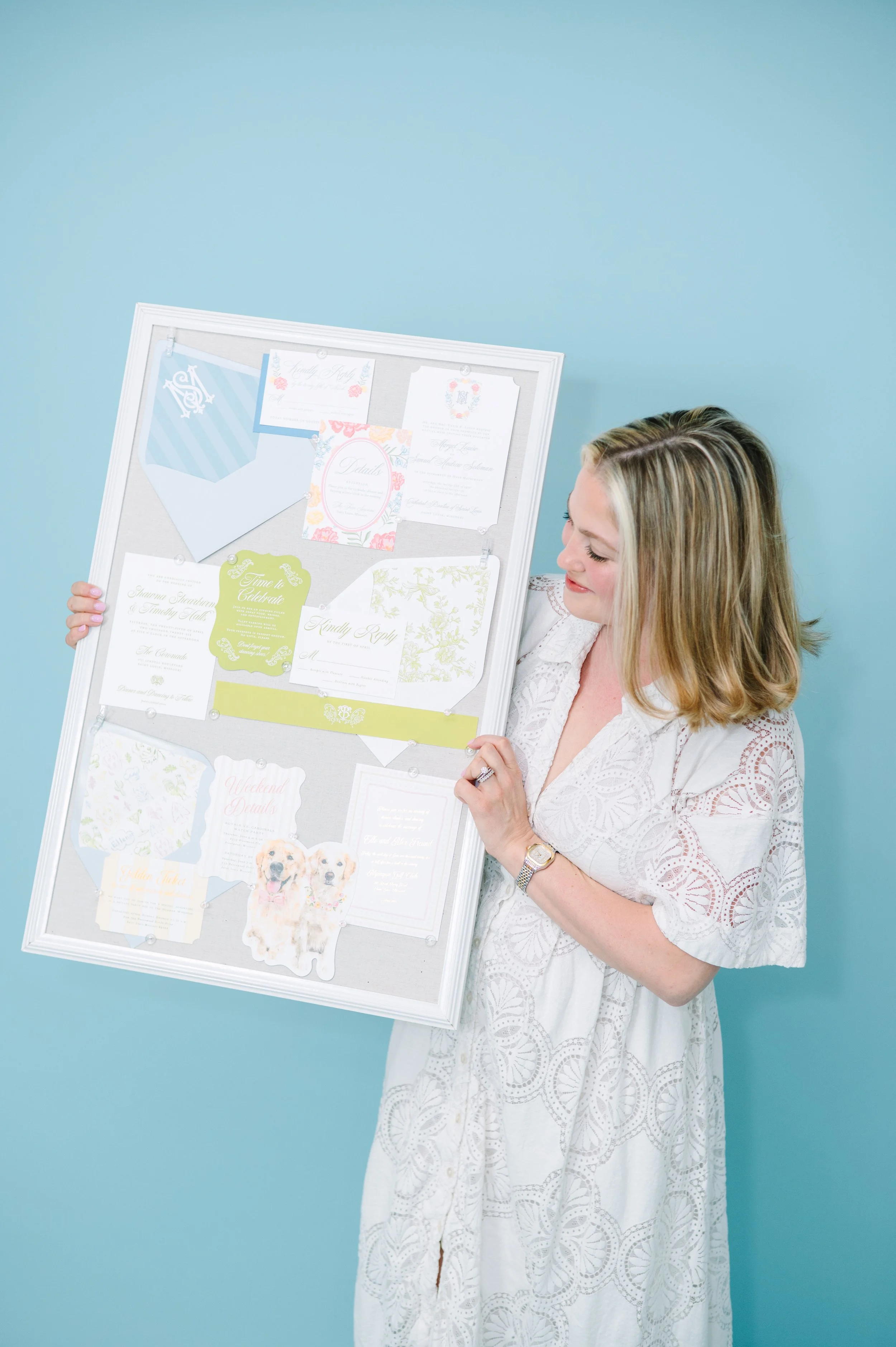 Woman in white dress looking at a large display board with invitations, cards, and photos against a light blue background.