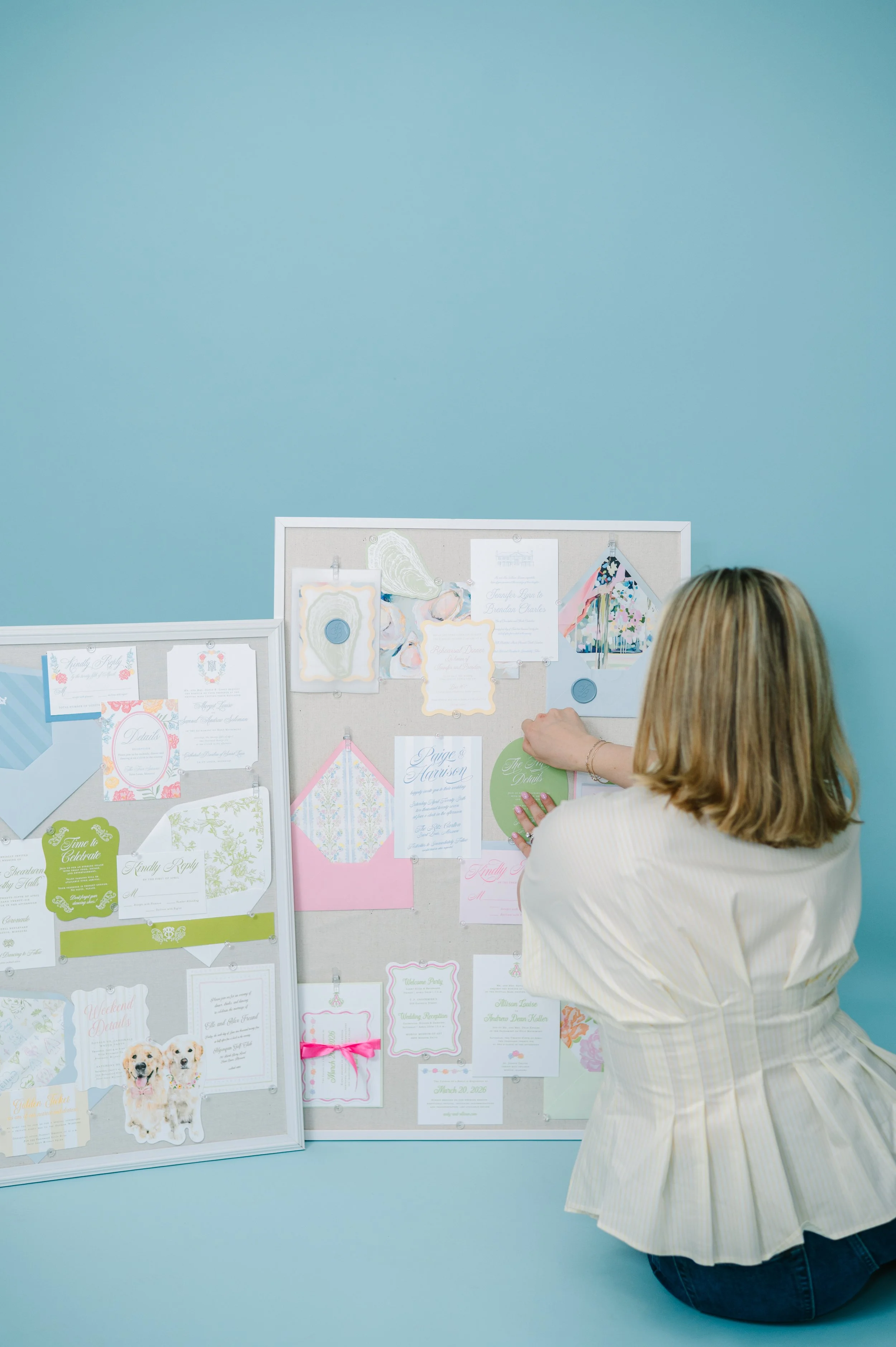 A woman kneeling in front of a bulletin board filled with wedding invitations and cards, against a light blue wall.