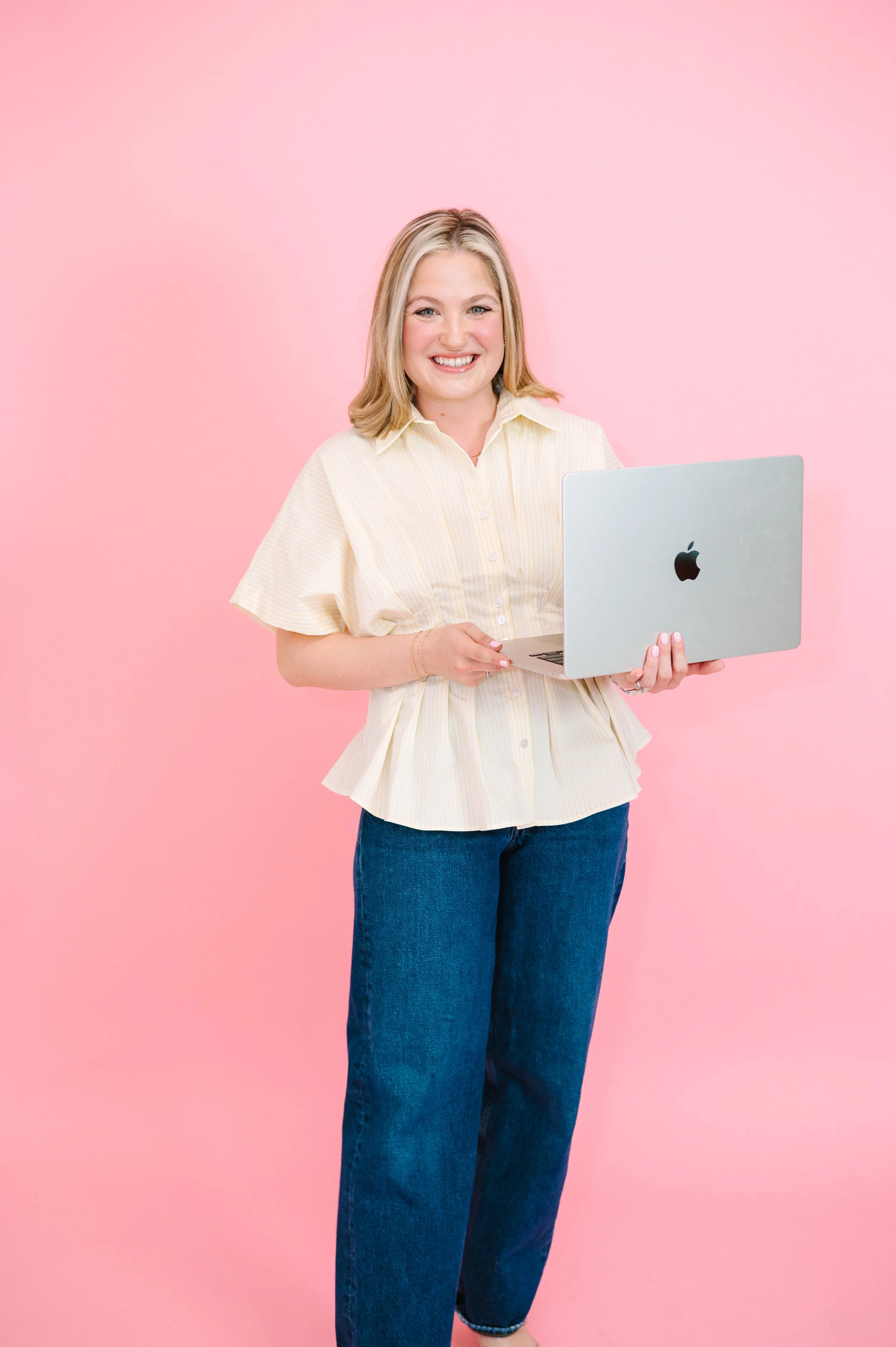 A woman with blonde hair smiling and holding a silver laptop in front of a pink background, dressed in a light yellow striped shirt and blue jeans.