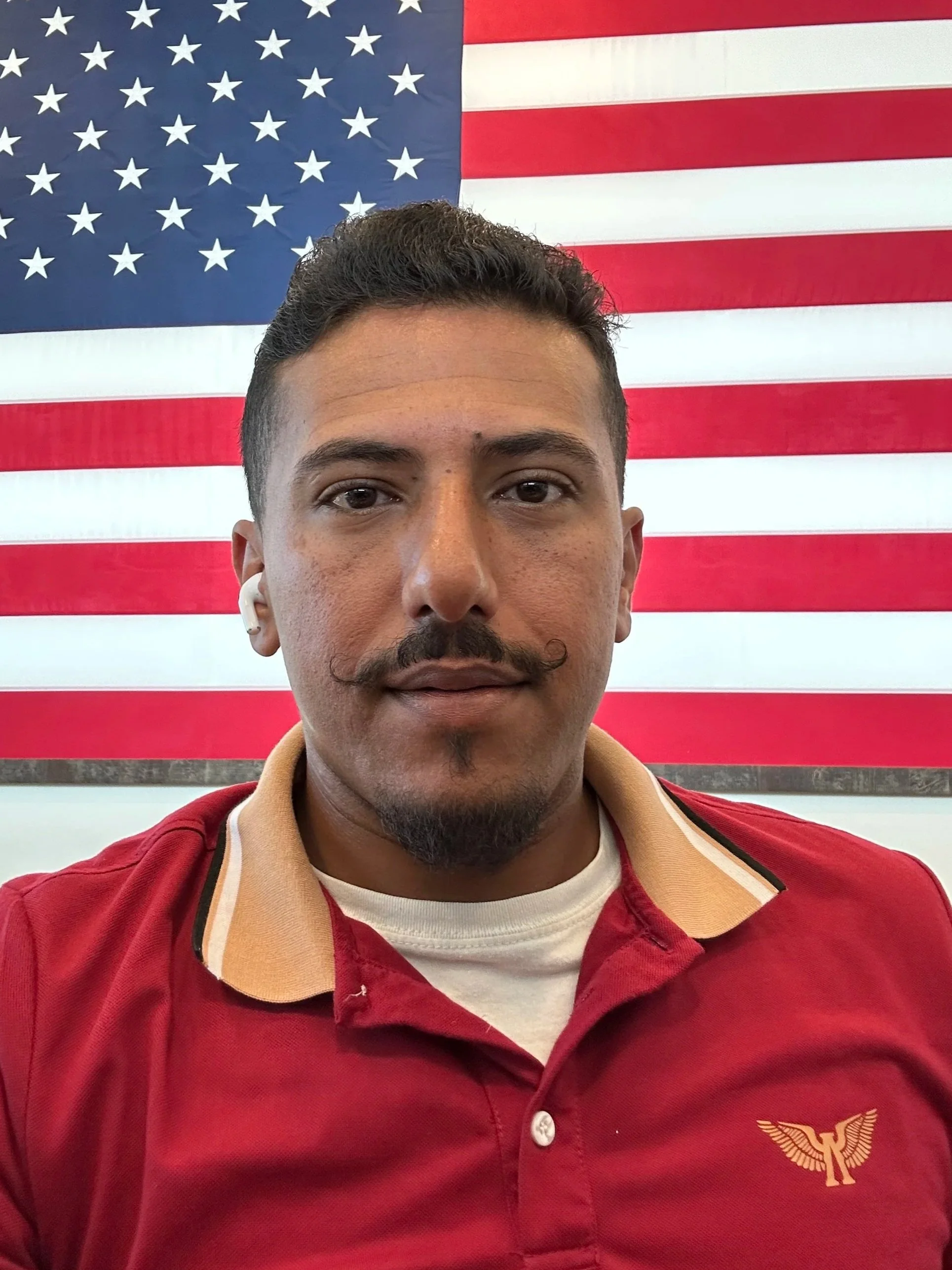 A man with short dark hair and a beard and mustache, wearing a red collared shirt with a beige trim, standing in front of an American flag.